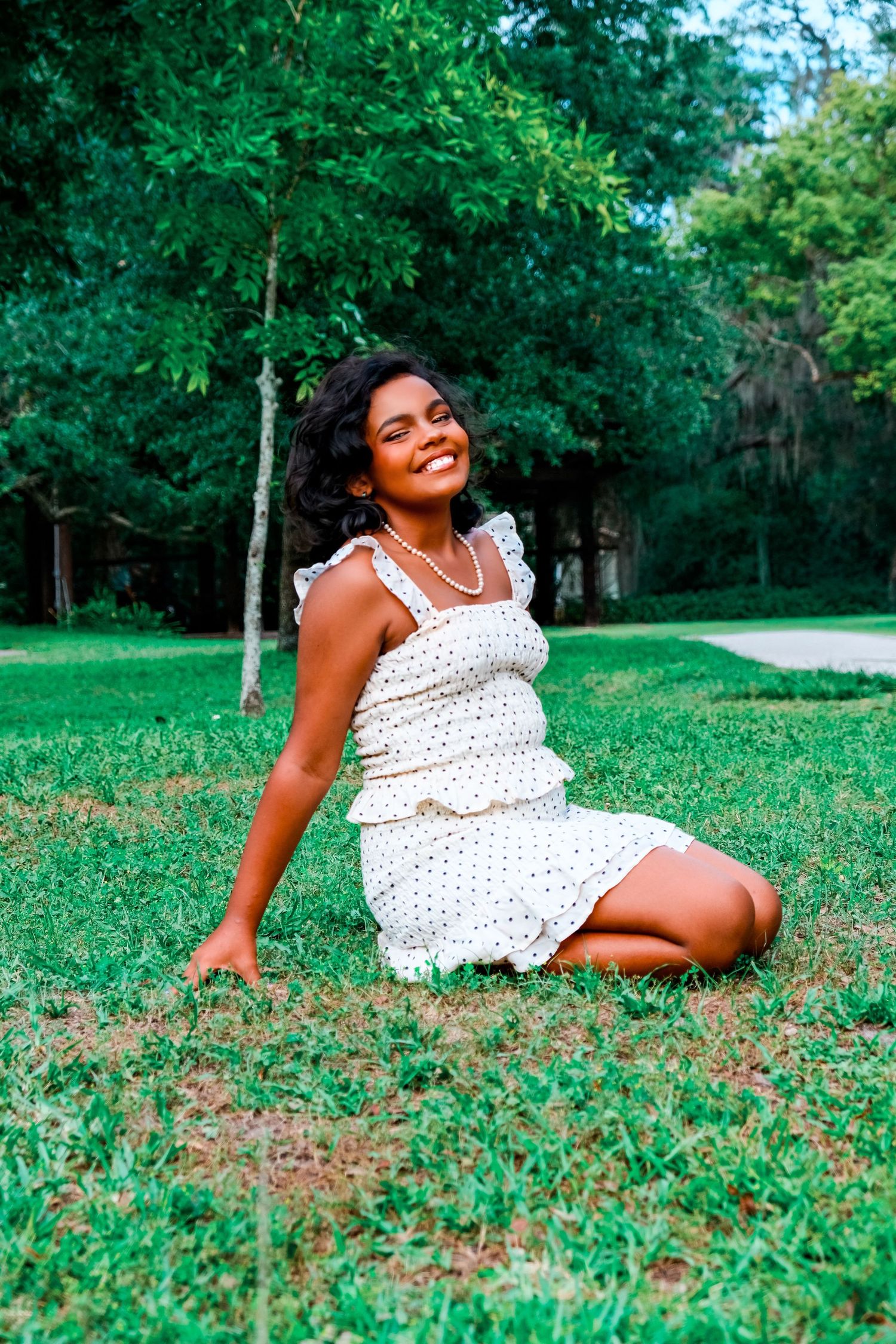 Smiling young woman in white polka dot dress sitting on green grass in a park surrounded by trees.