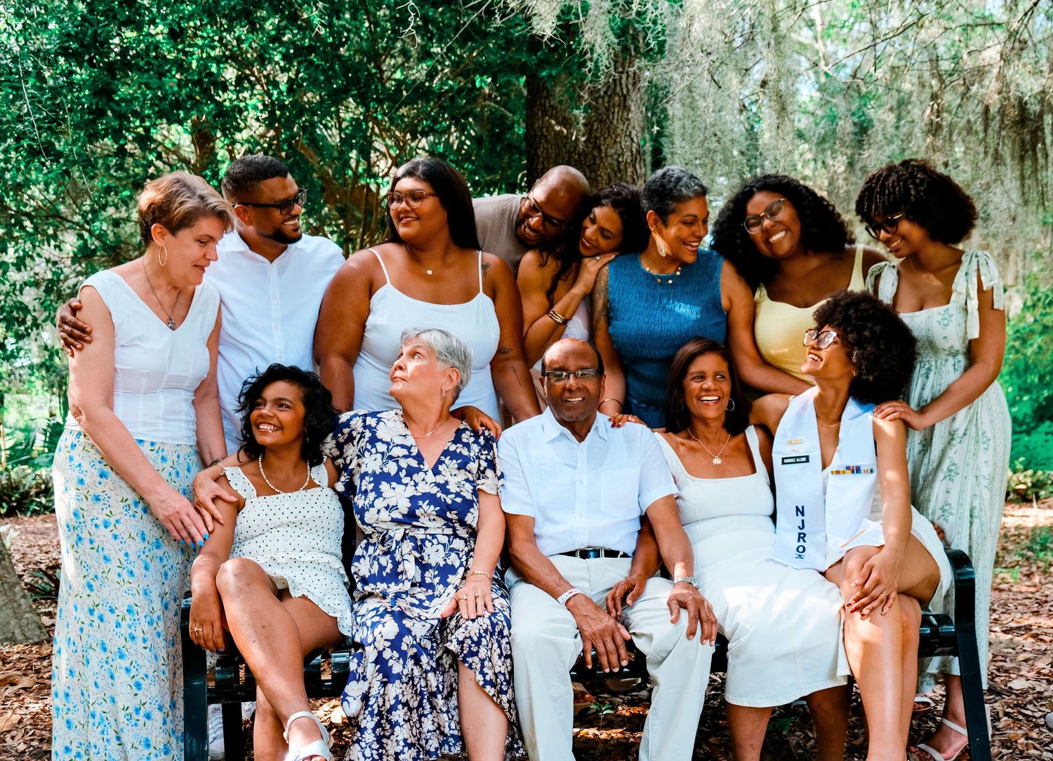 Large diverse family group smiling together outdoors under moss-covered trees, dressed in white and blue outfits.