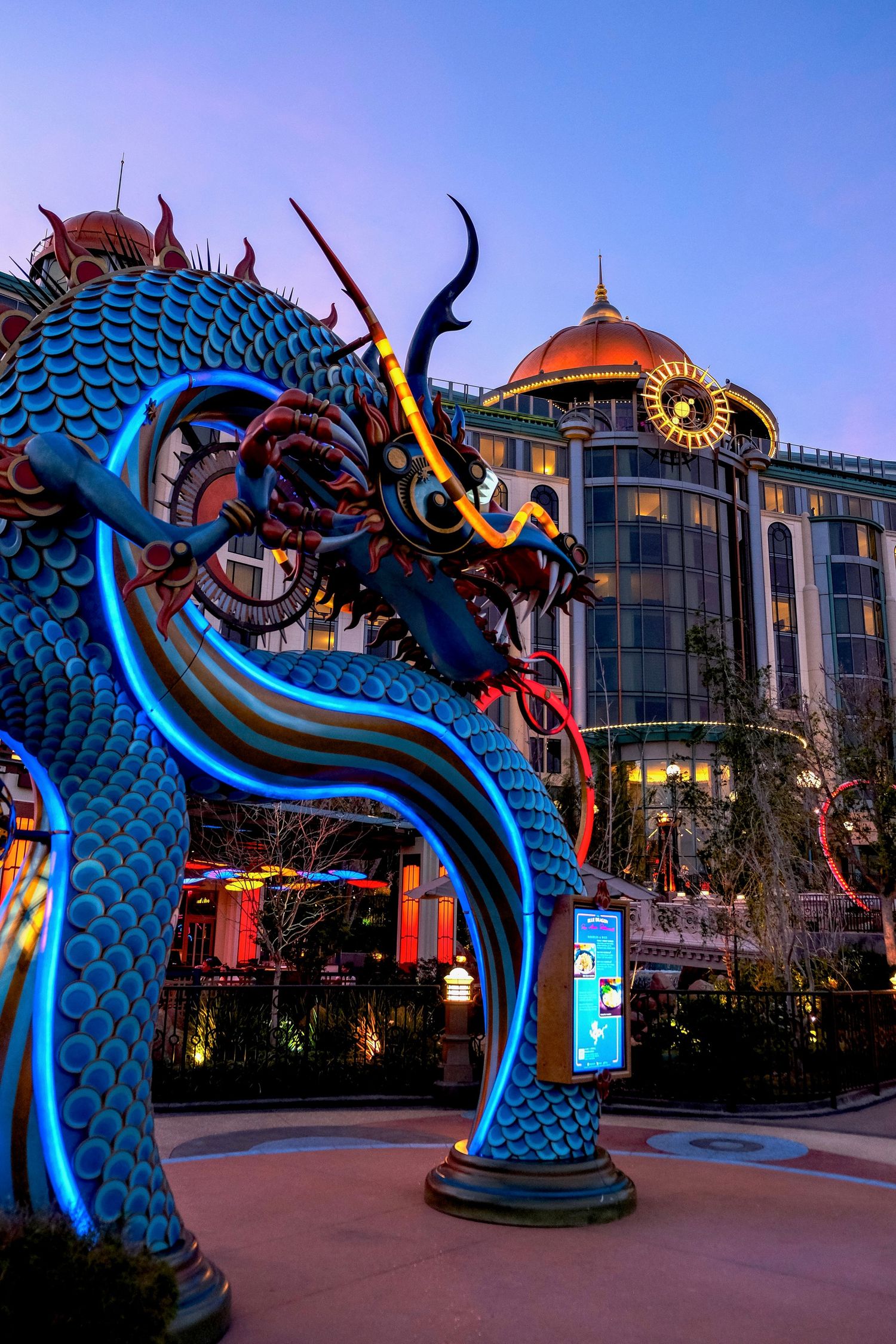 Illuminated blue dragon sculpture at dusk with ornate Asian-style building featuring copper dome in background.