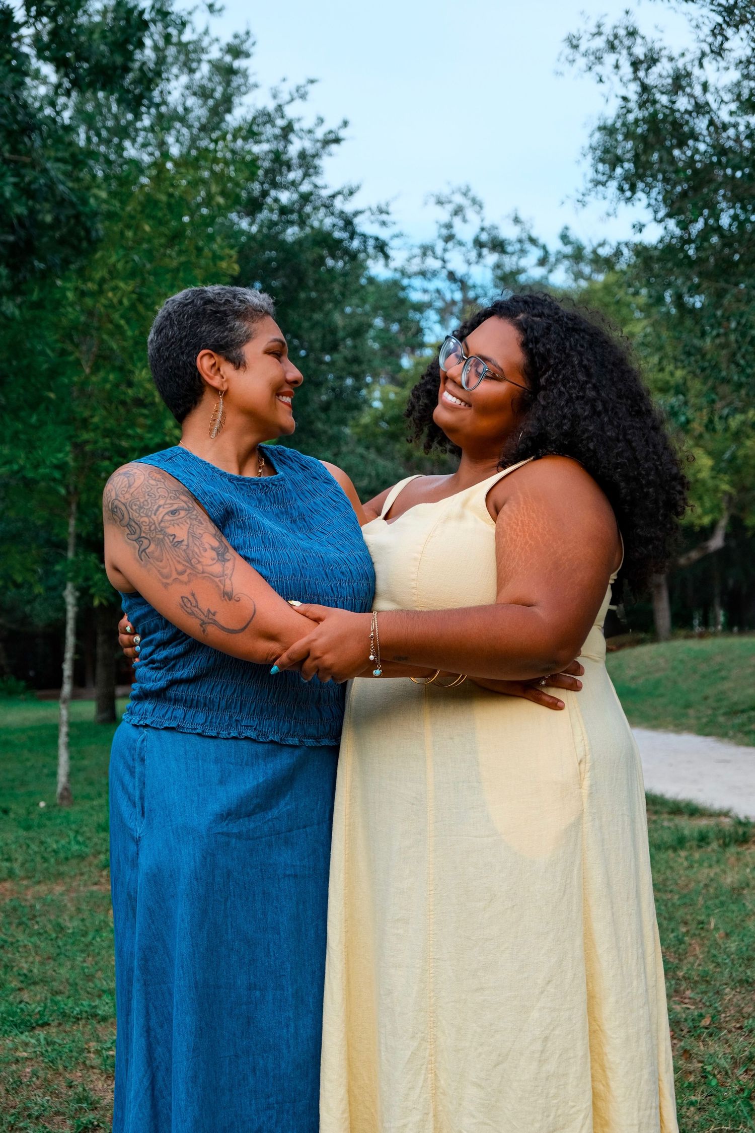 Two Black women embracing and smiling at each other in a park, one in blue denim dress, one in yellow gown.