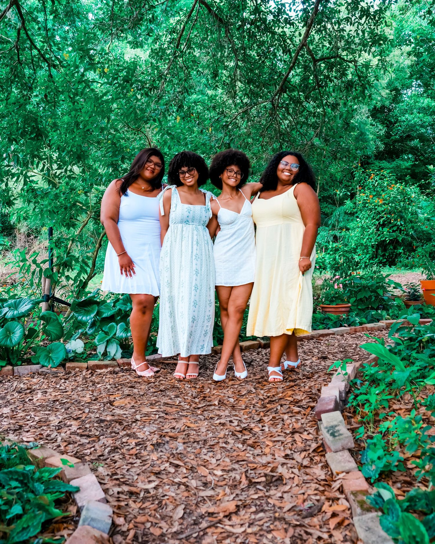 Four Black women in pastel summer dresses posing together in a lush green garden with mulch pathway.