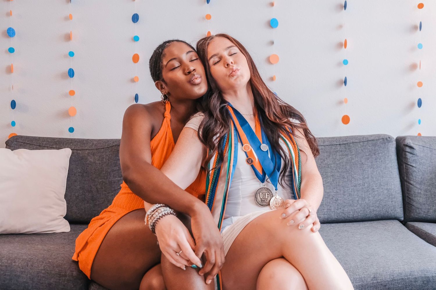 Two young women sitting on a gray couch making kissy faces, one wearing medals and colorful party decorations in background.