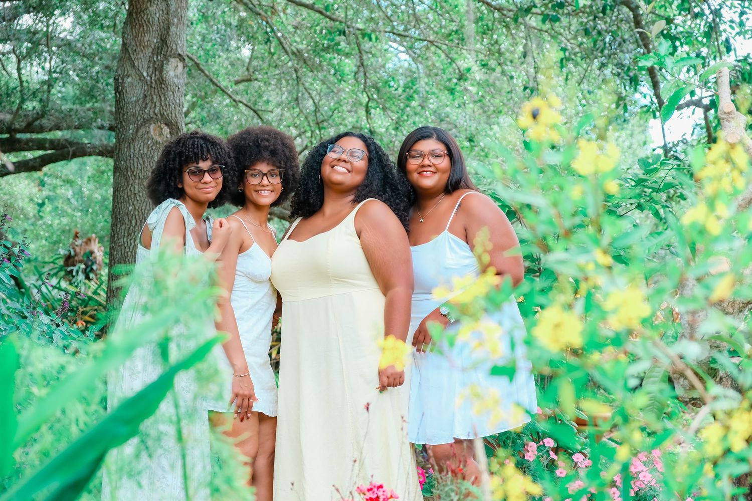 Four Black women in white dresses smiling together outdoors among lush green trees and yellow flowers.