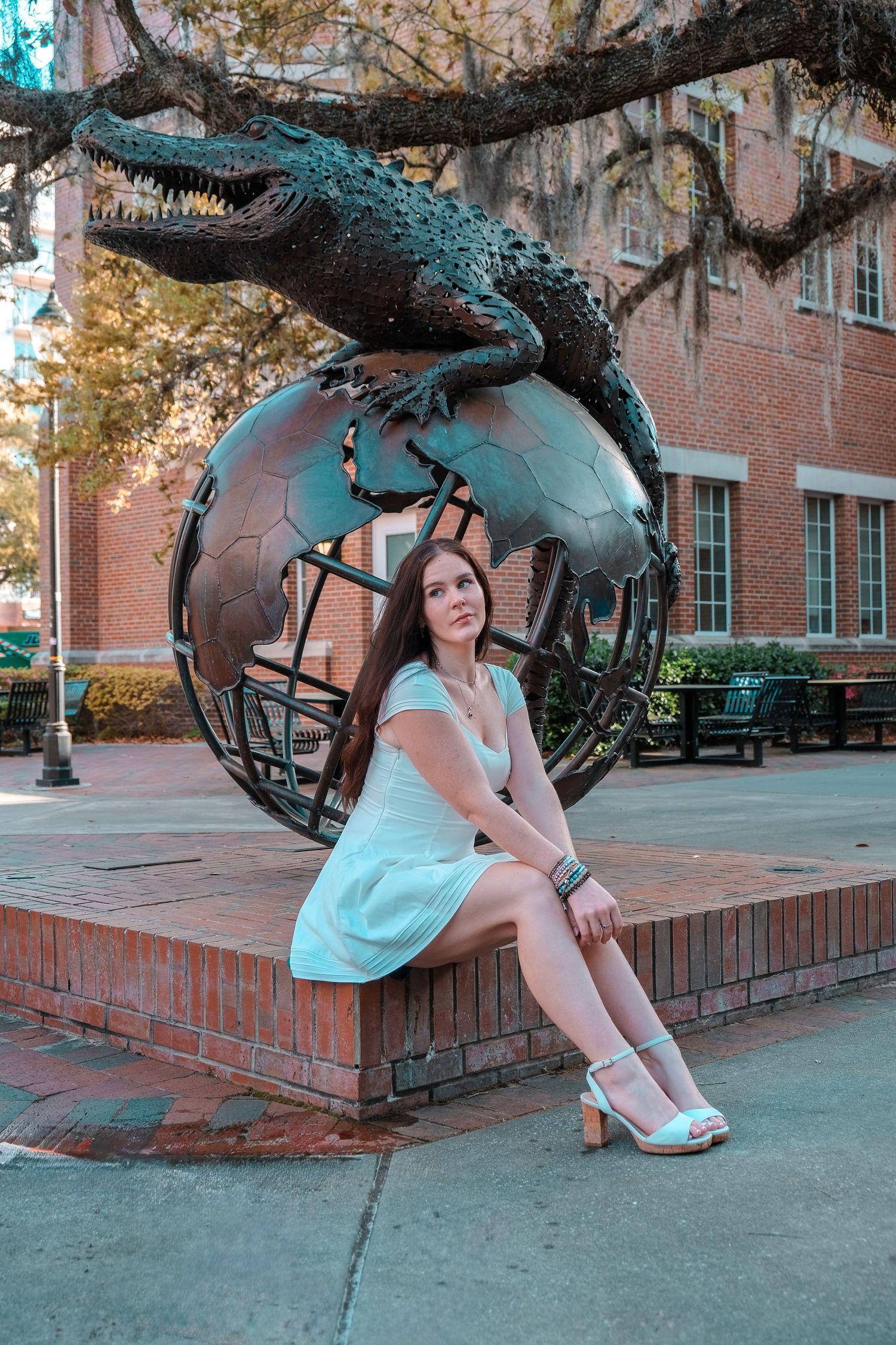 Young woman in light blue dress sits beside bronze alligator globe sculpture at University of Florida.