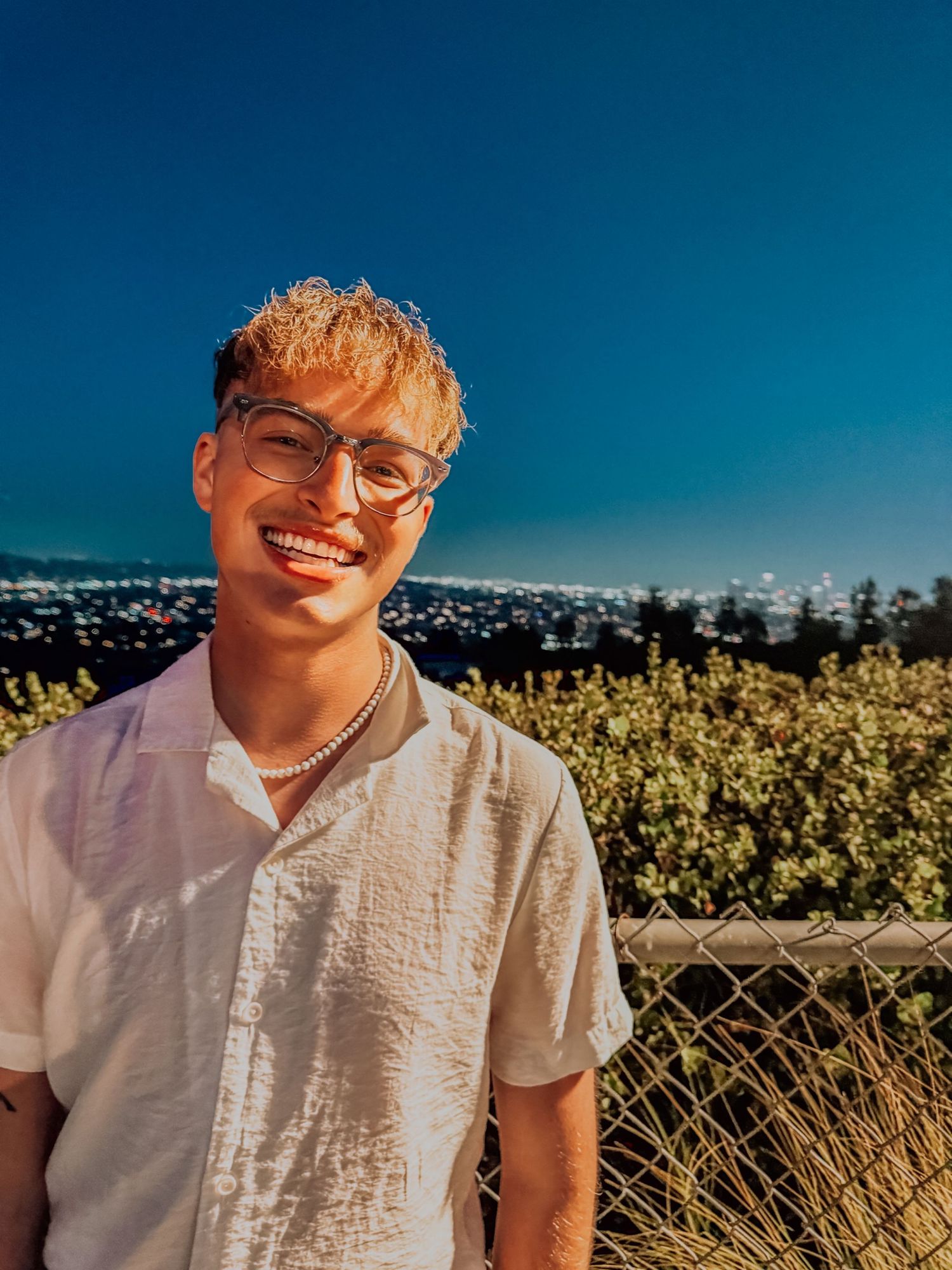 Young man with blonde highlights and glasses smiling at sunset with city lights in the background.