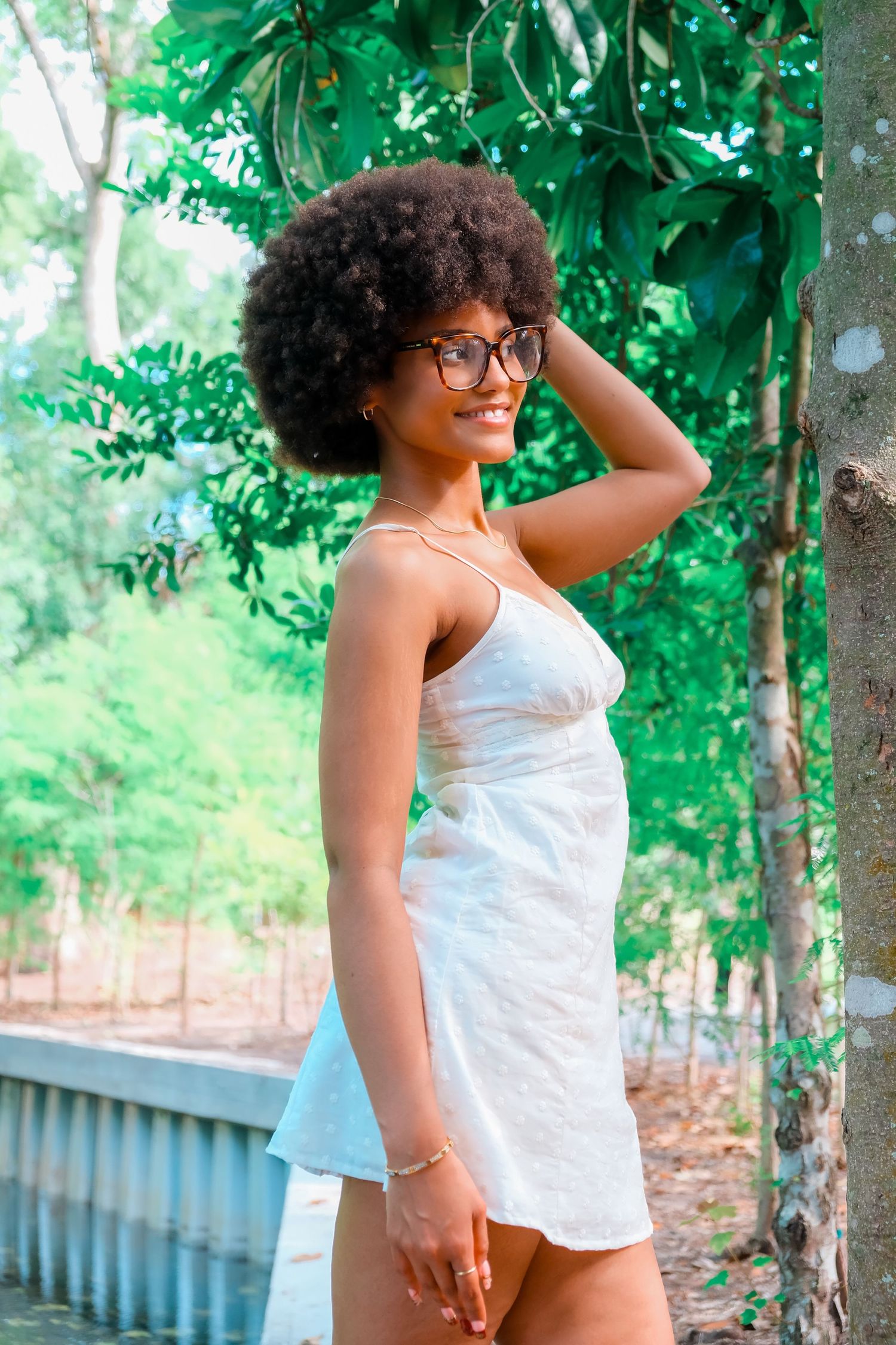 Young Black woman with afro hair and glasses wearing a white mini dress, smiling outdoors among green trees.