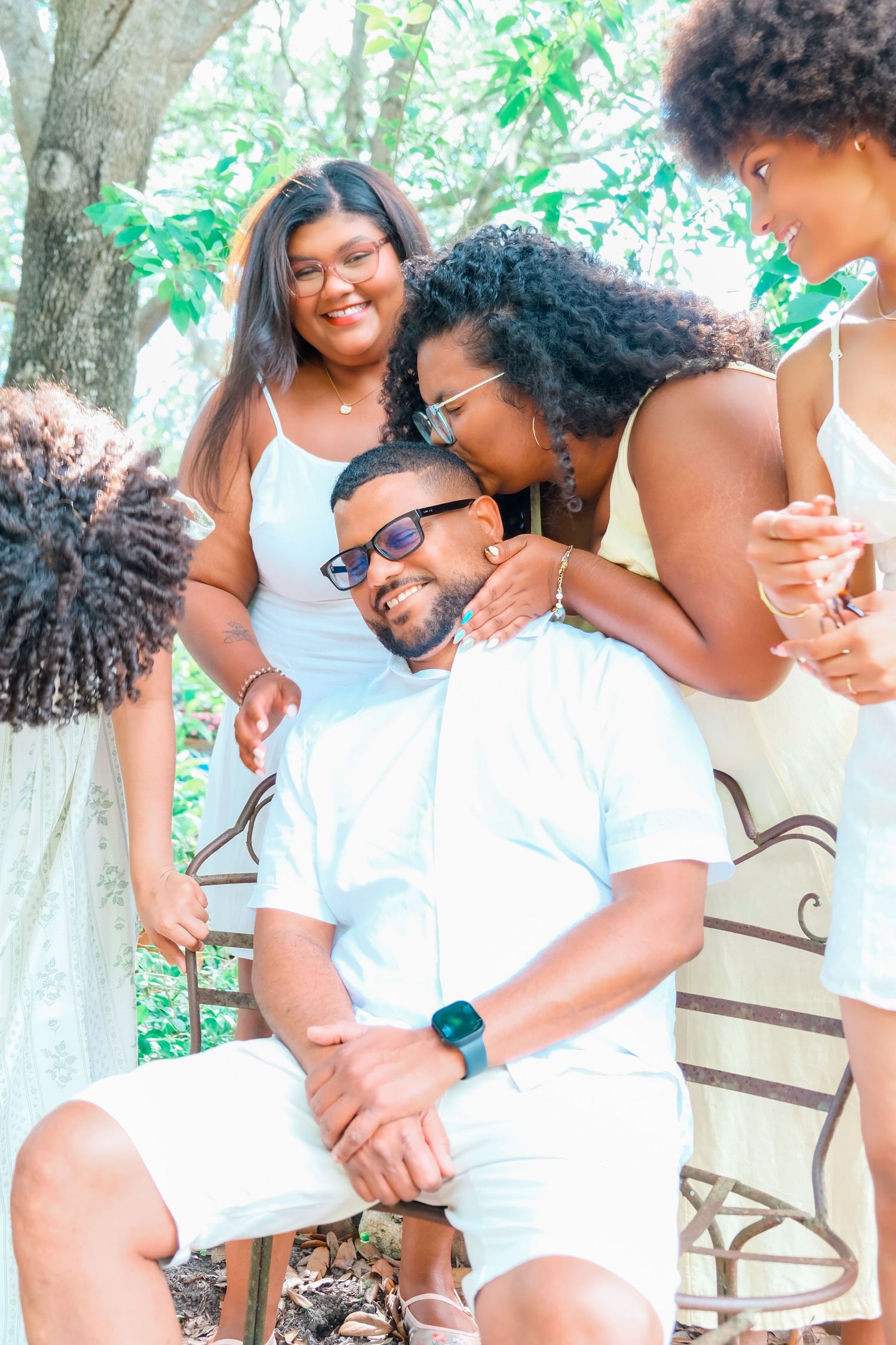 Group of friends in white outfits gather outdoors, smiling and embracing near trees on a sunny day.