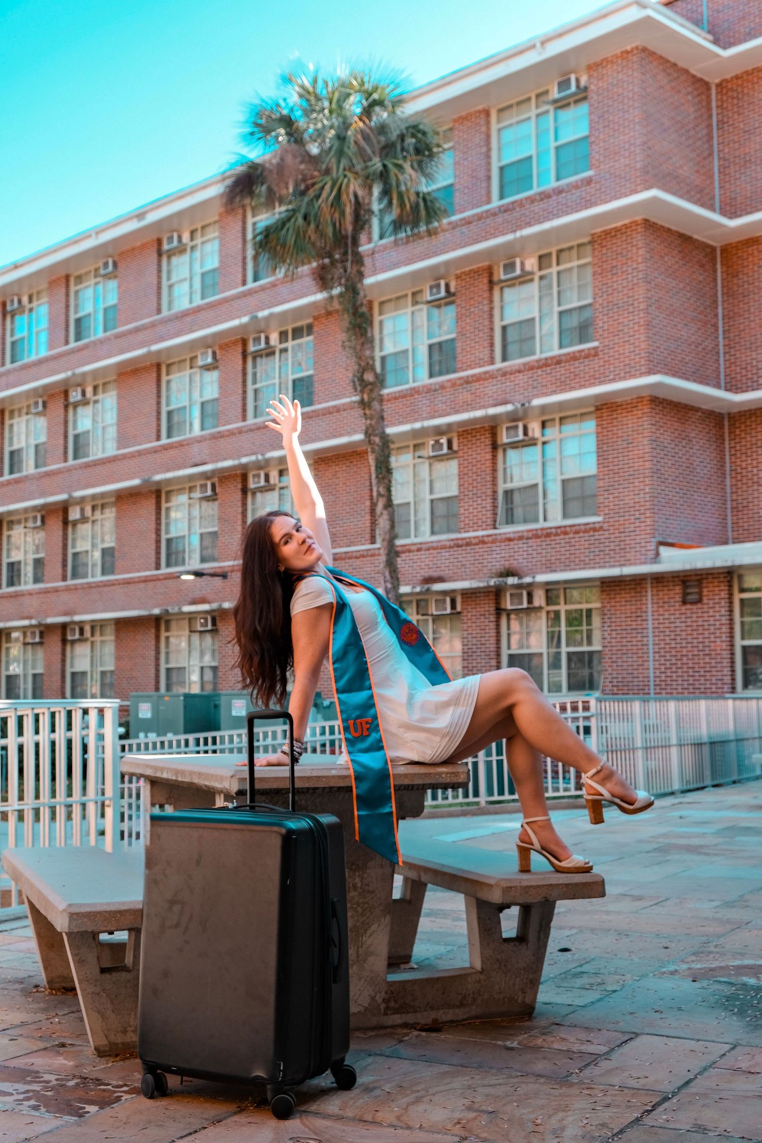 Excited female graduate in UF sash sits on bench with suitcase, arm raised, outside brick campus building.