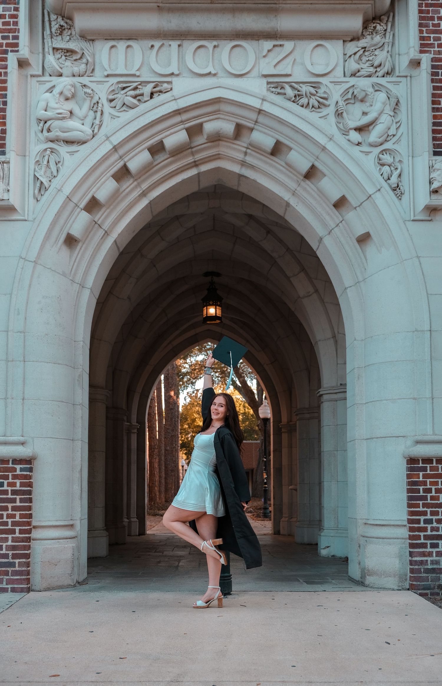 Young woman in mint dress and black gown poses under ornate stone arch at university building entrance.