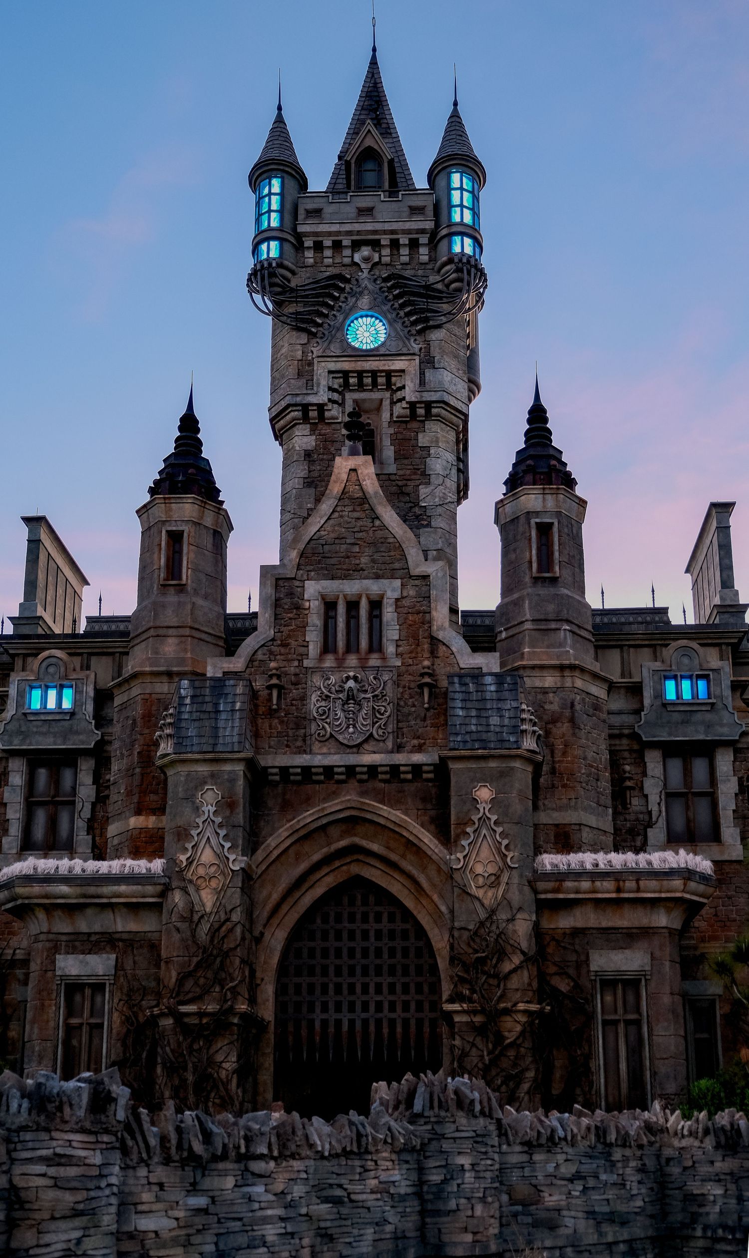 Gothic castle with glowing blue windows and clock tower at dusk, featuring ornate stone carvings and pointed spires.