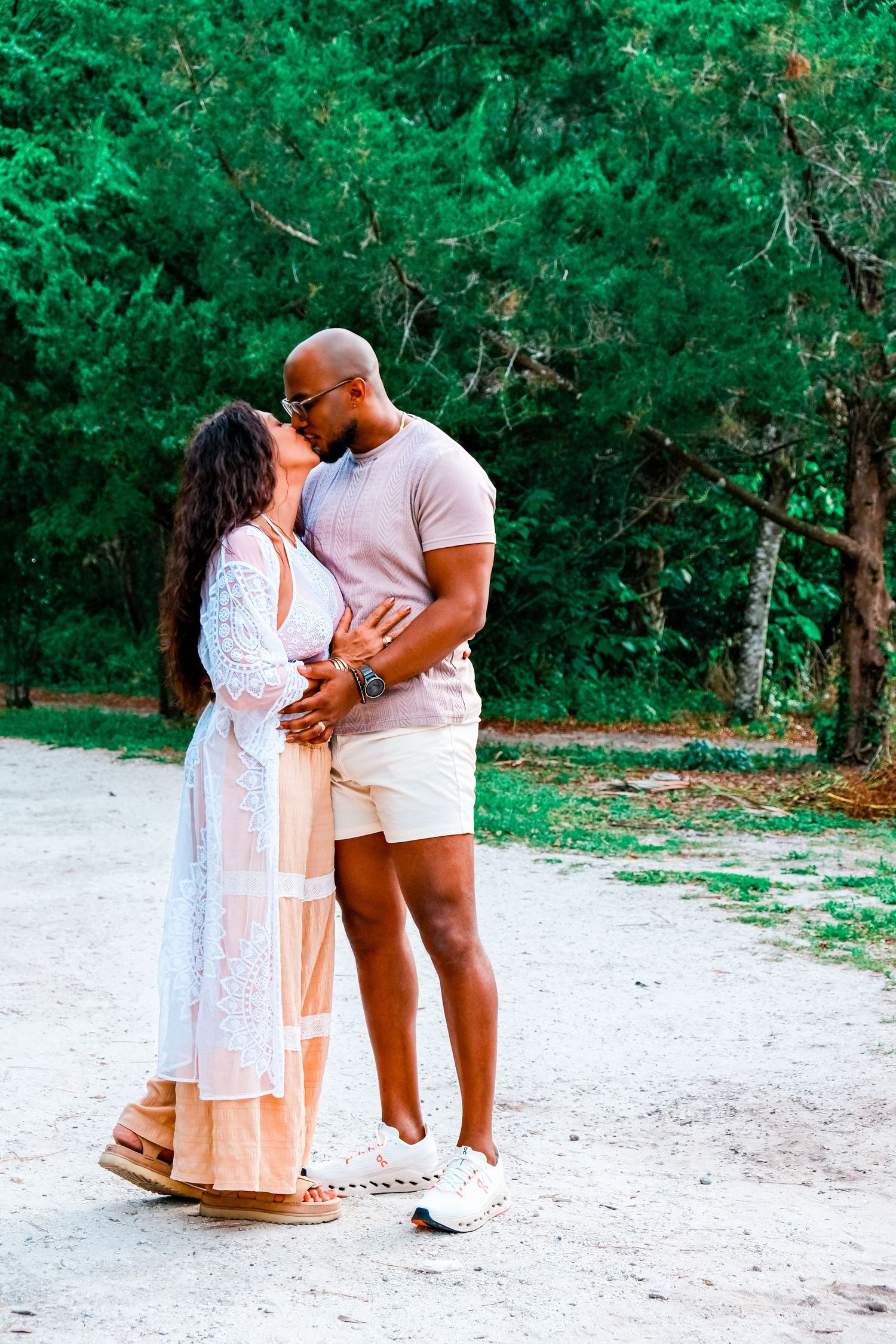 A couple shares a romantic kiss outdoors on a sandy path surrounded by lush green trees.