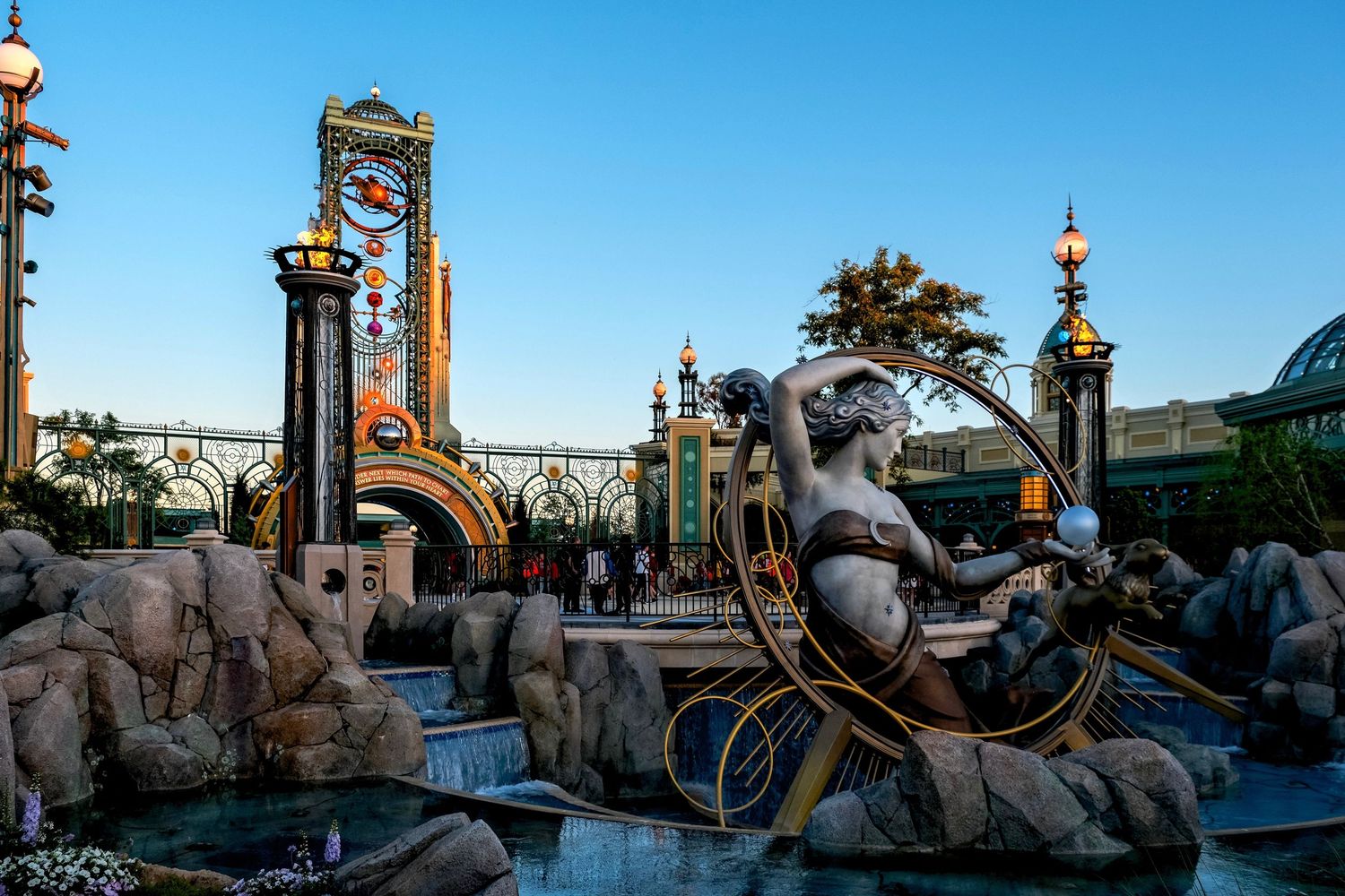 Bronze fountain statue at a fantasy theme park entrance with ornate golden gates and clock tower at sunset.