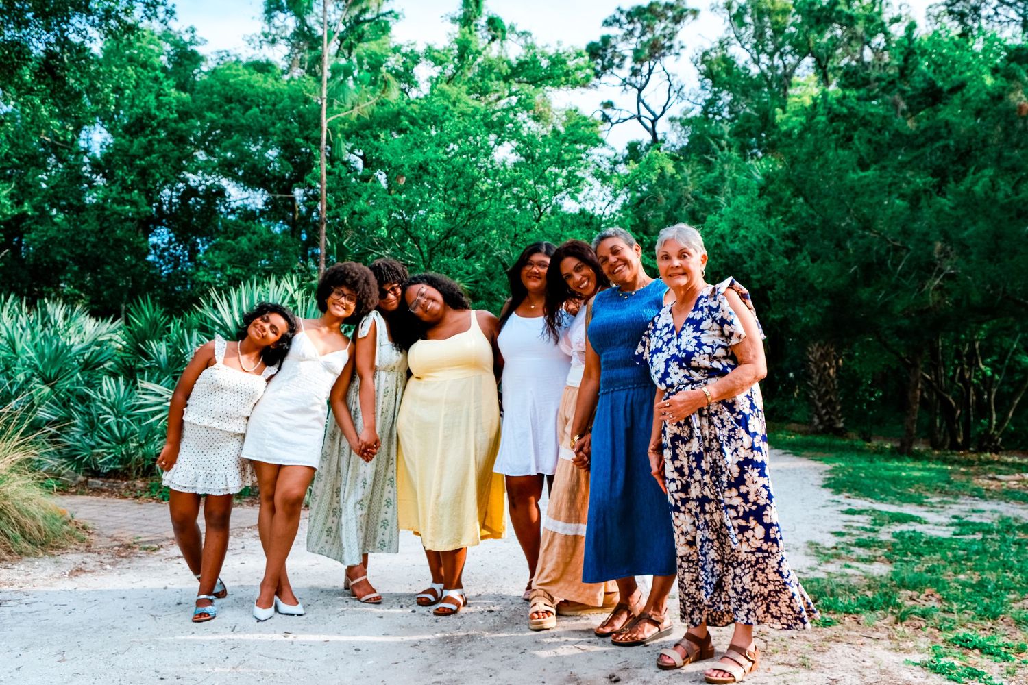 Group of women in summer dresses smiling together outdoors on a path surrounded by lush green trees.