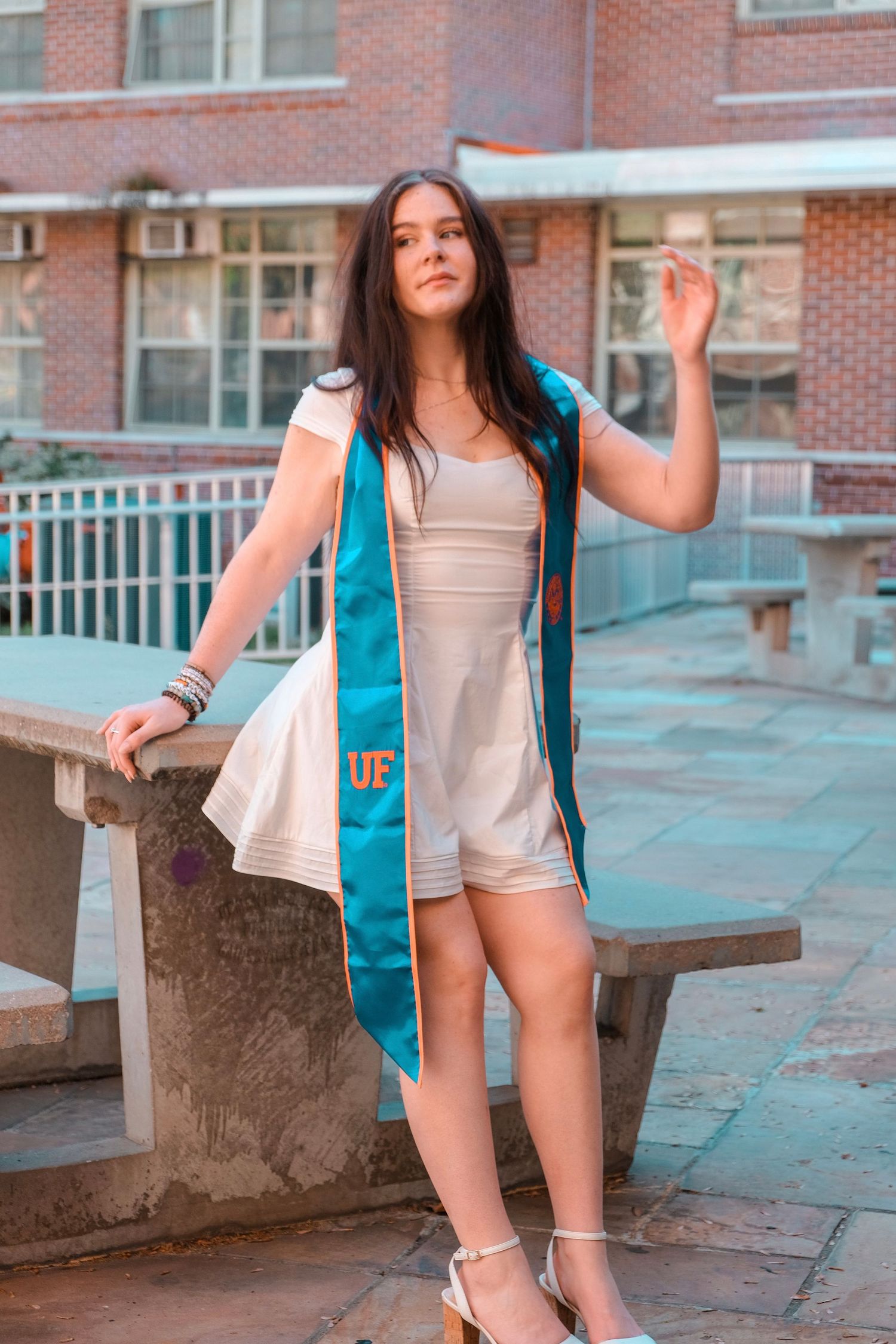 Female graduate wearing UF teal sash over white dress poses confidently outside brick university building.
