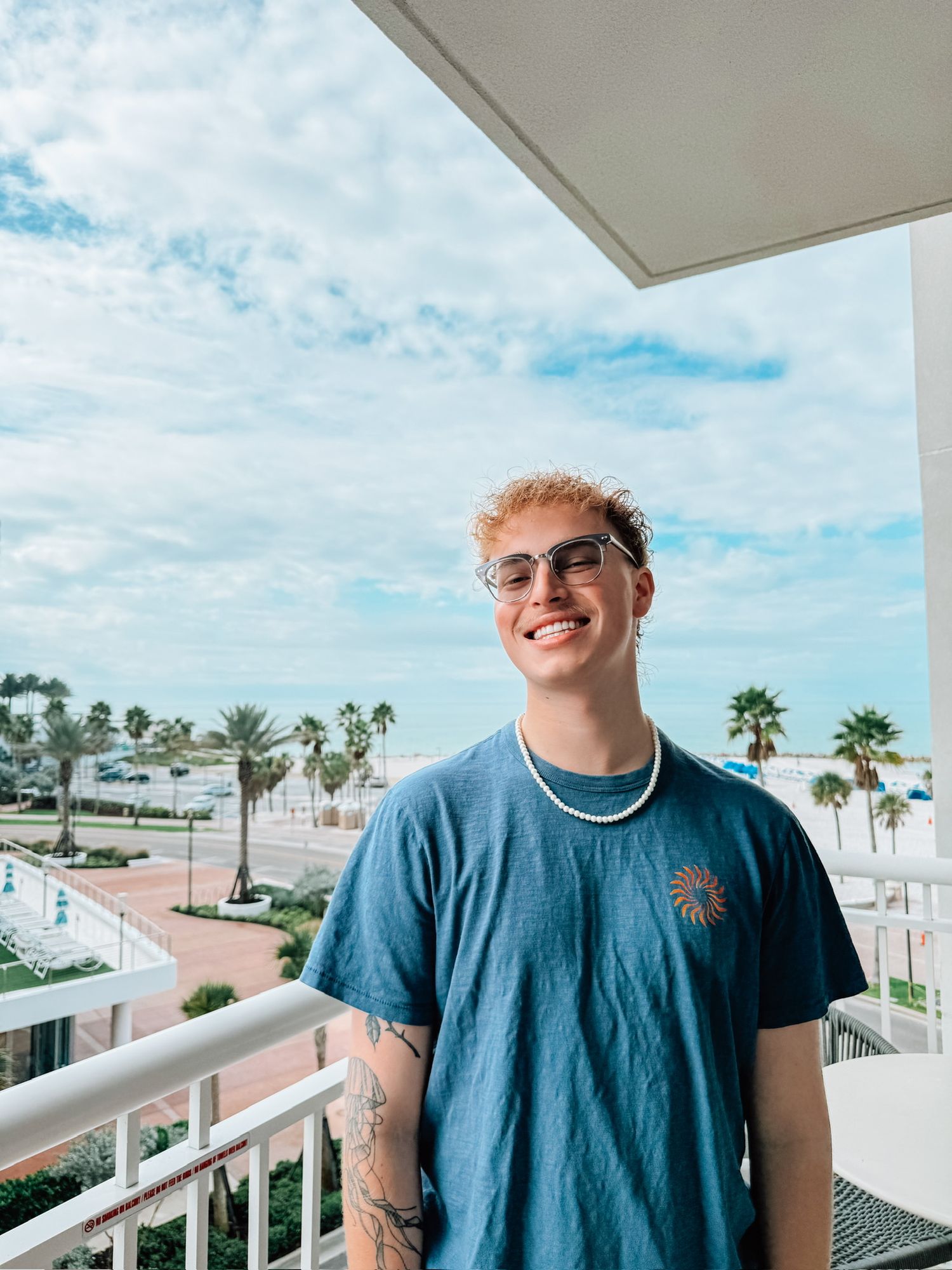 A person wearing a blue t-shirt stands on a balcony with palm trees and white buildings visible in the background.