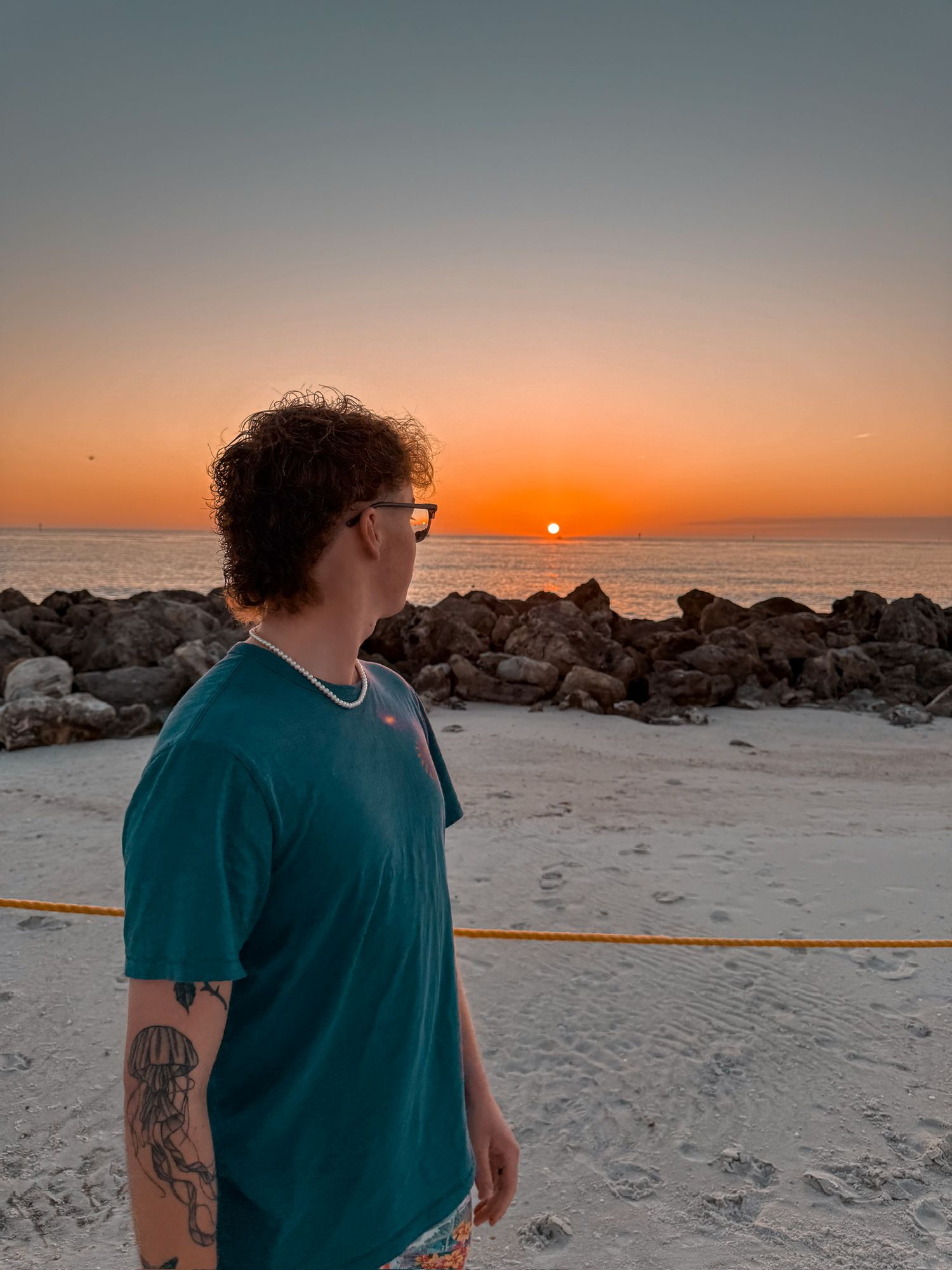 A person in a teal shirt stands on a beach at sunset with rocky jetty and orange sky in the background.