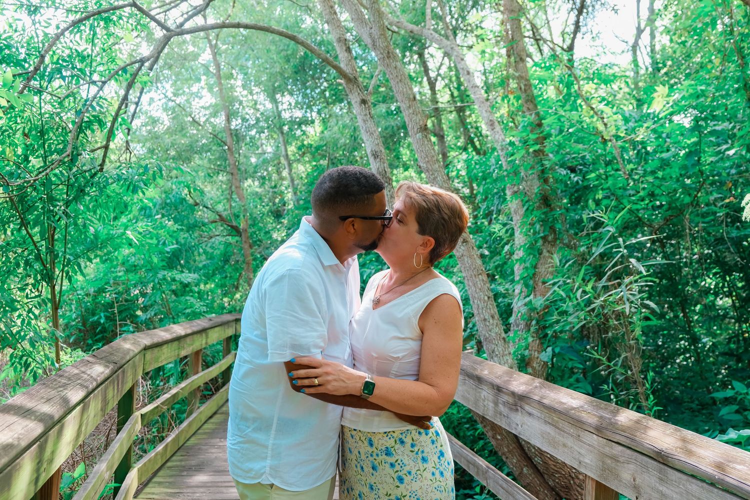 Couple sharing a romantic kiss on a wooden bridge surrounded by lush green trees outdoors.