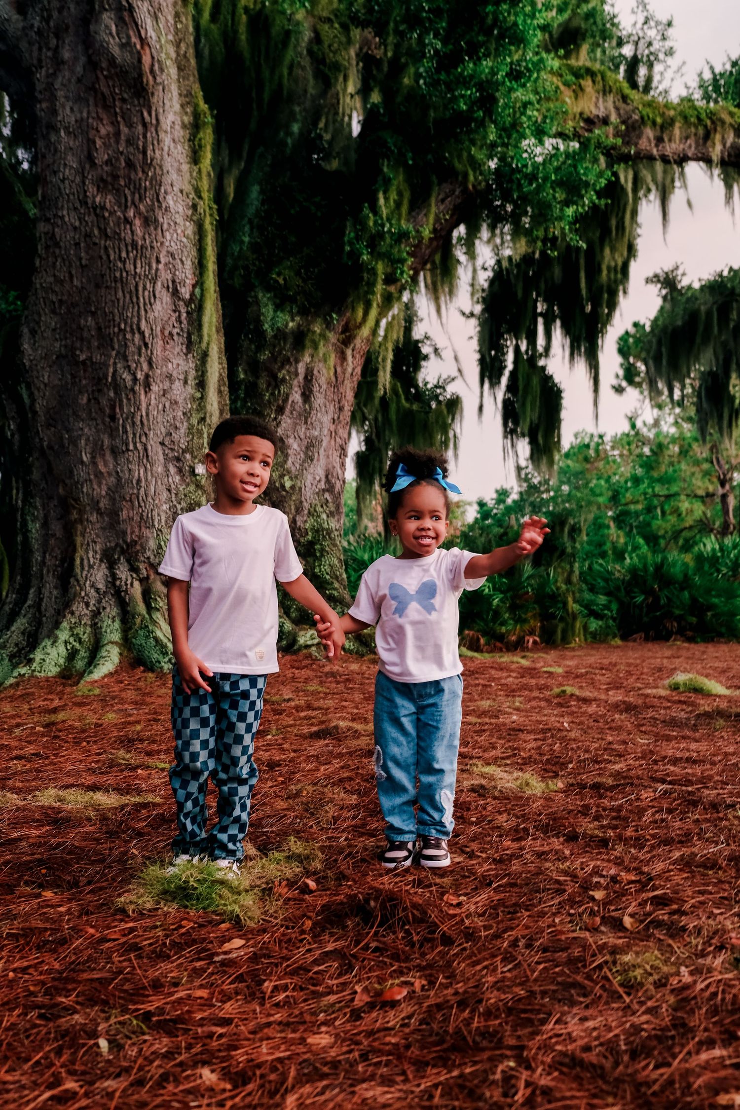 Two young children holding hands near a large moss-draped tree on a red mulch path outdoors.