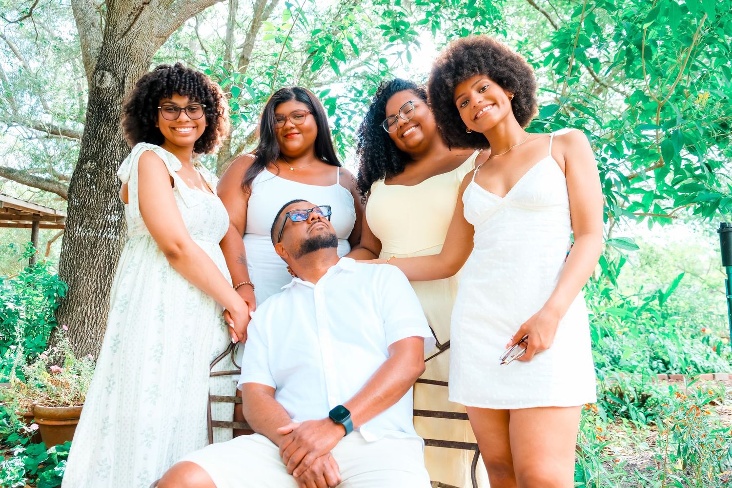 Black father seated outdoors surrounded by three daughters in white and neutral outfits among green trees.