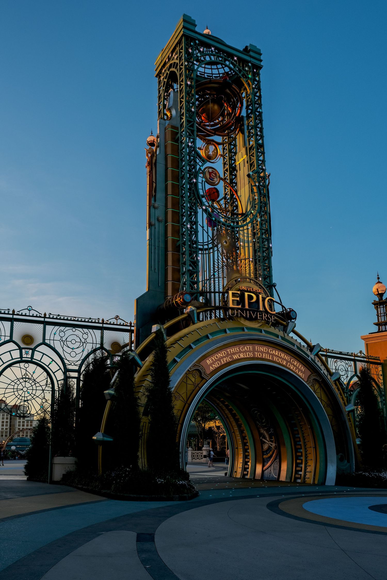 Grand entrance tower of Epic Universe theme park at dusk, featuring ornate metalwork and glowing signage against a blue sky.