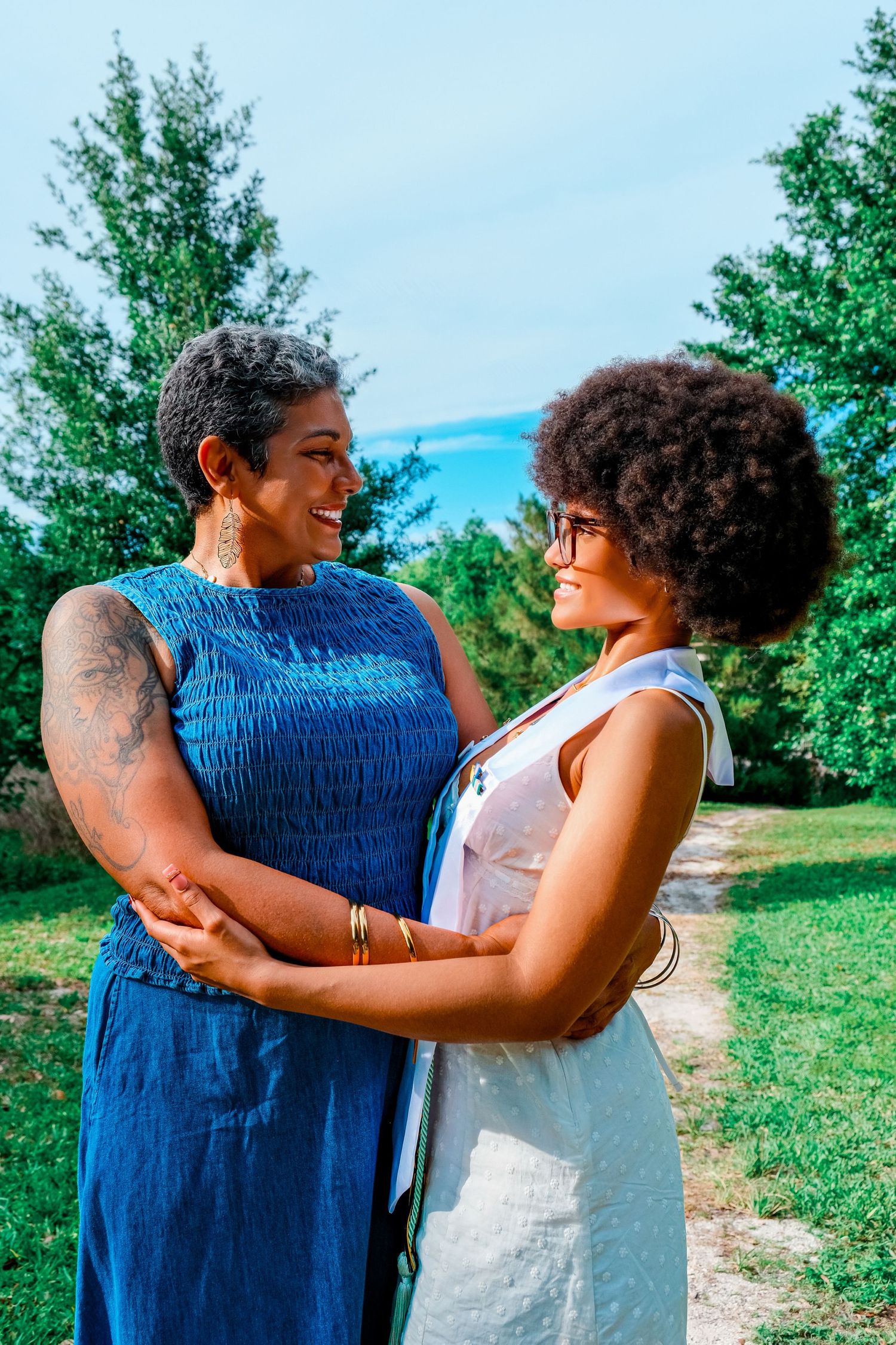 Two Black women holding hands and smiling at each other outdoors, one in blue dress, one in white dress.