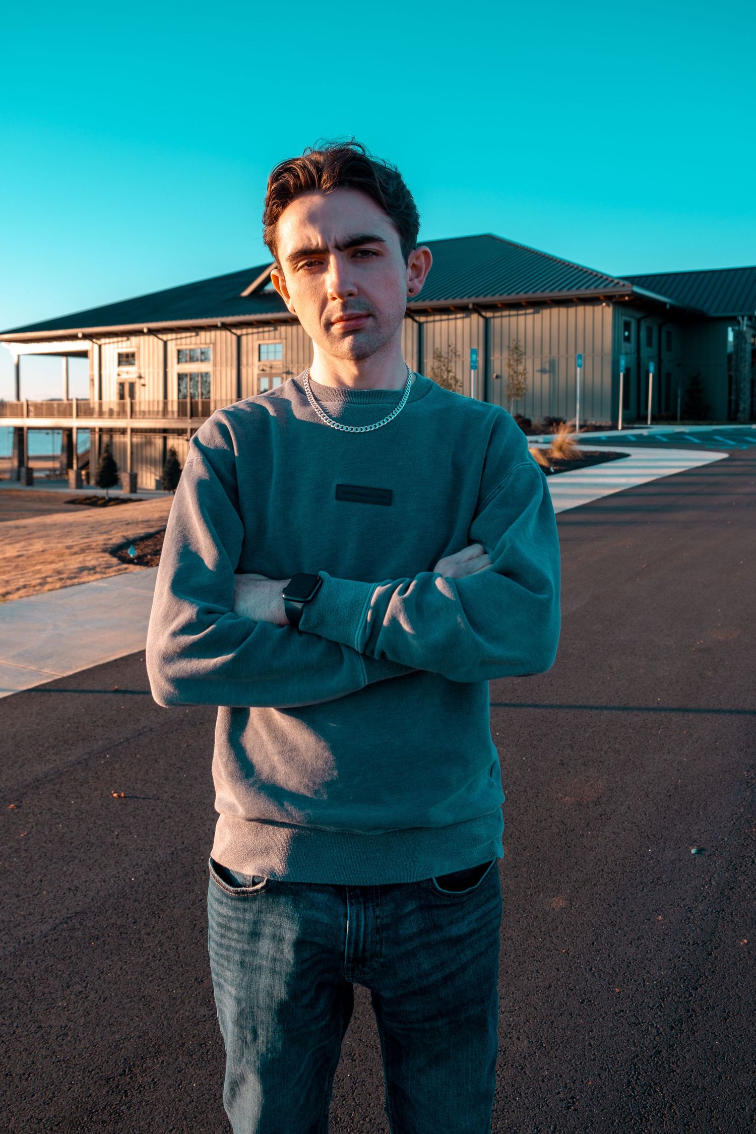 Young man in teal sweatshirt stands arms crossed outdoors near modern building under clear blue sky.