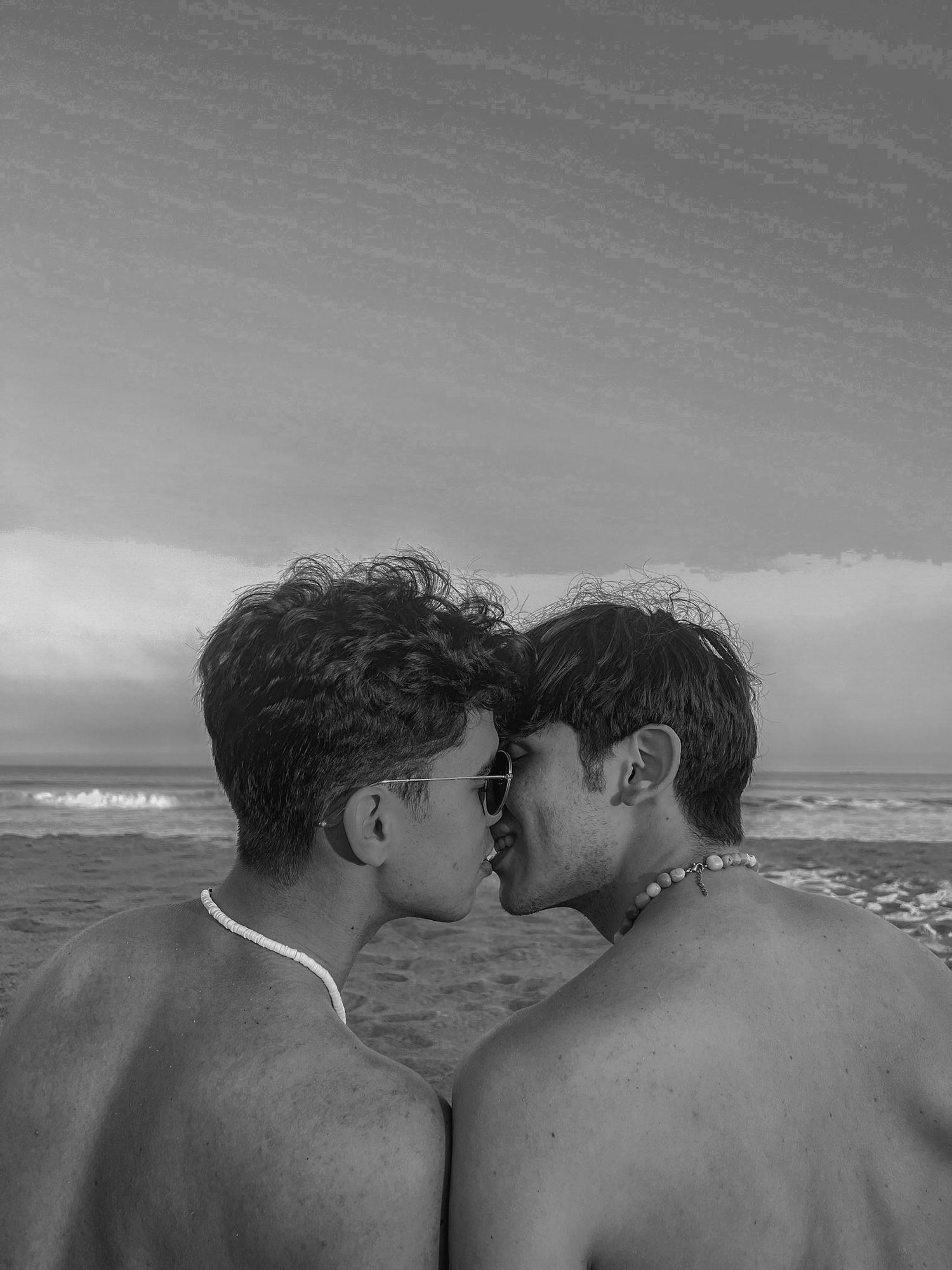 Two shirtless young men sharing a kiss on a beach in a black and white photo.