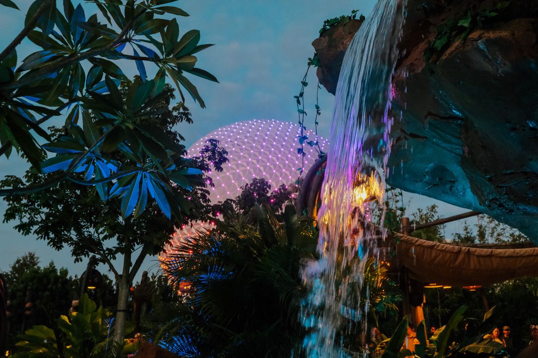 Epcot's illuminated geodesic sphere rises behind tropical foliage and a colorful waterfall at dusk in Walt Disney World.