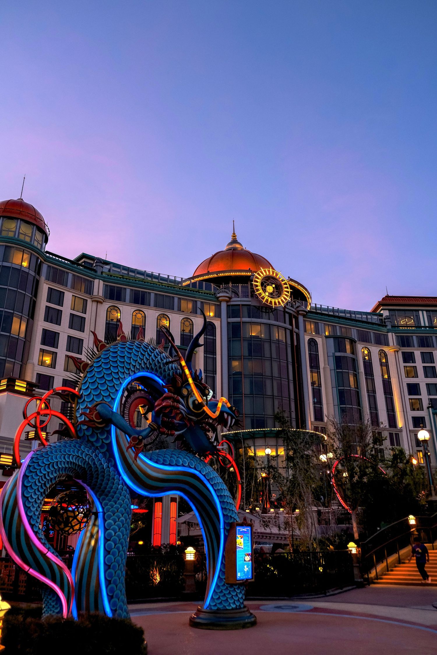 Illuminated dragon sculpture at dusk in front of a grand hotel with copper dome and ornate architecture.
