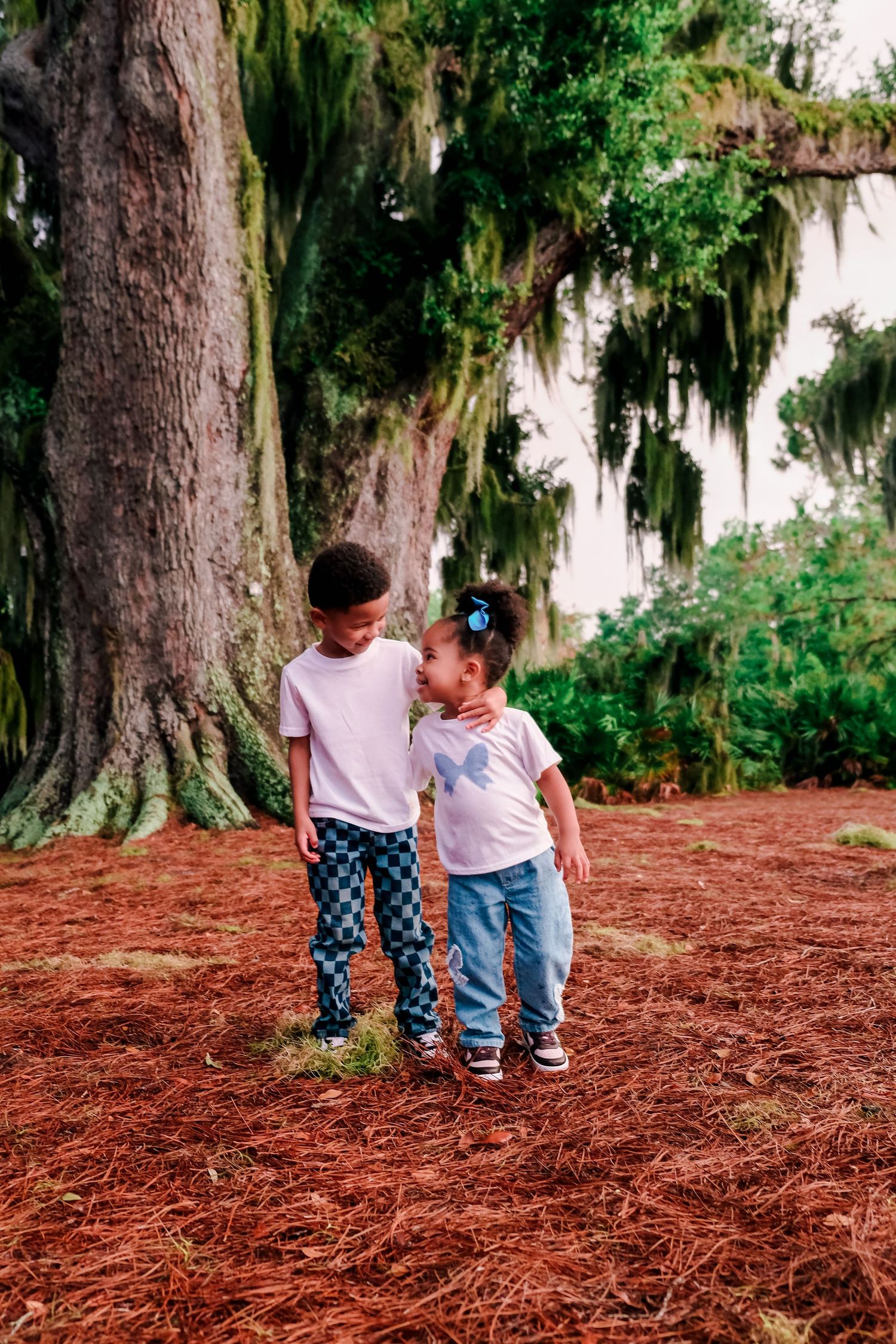 Two children stand together under a large moss-covered tree on a bed of red pine needles outdoors.