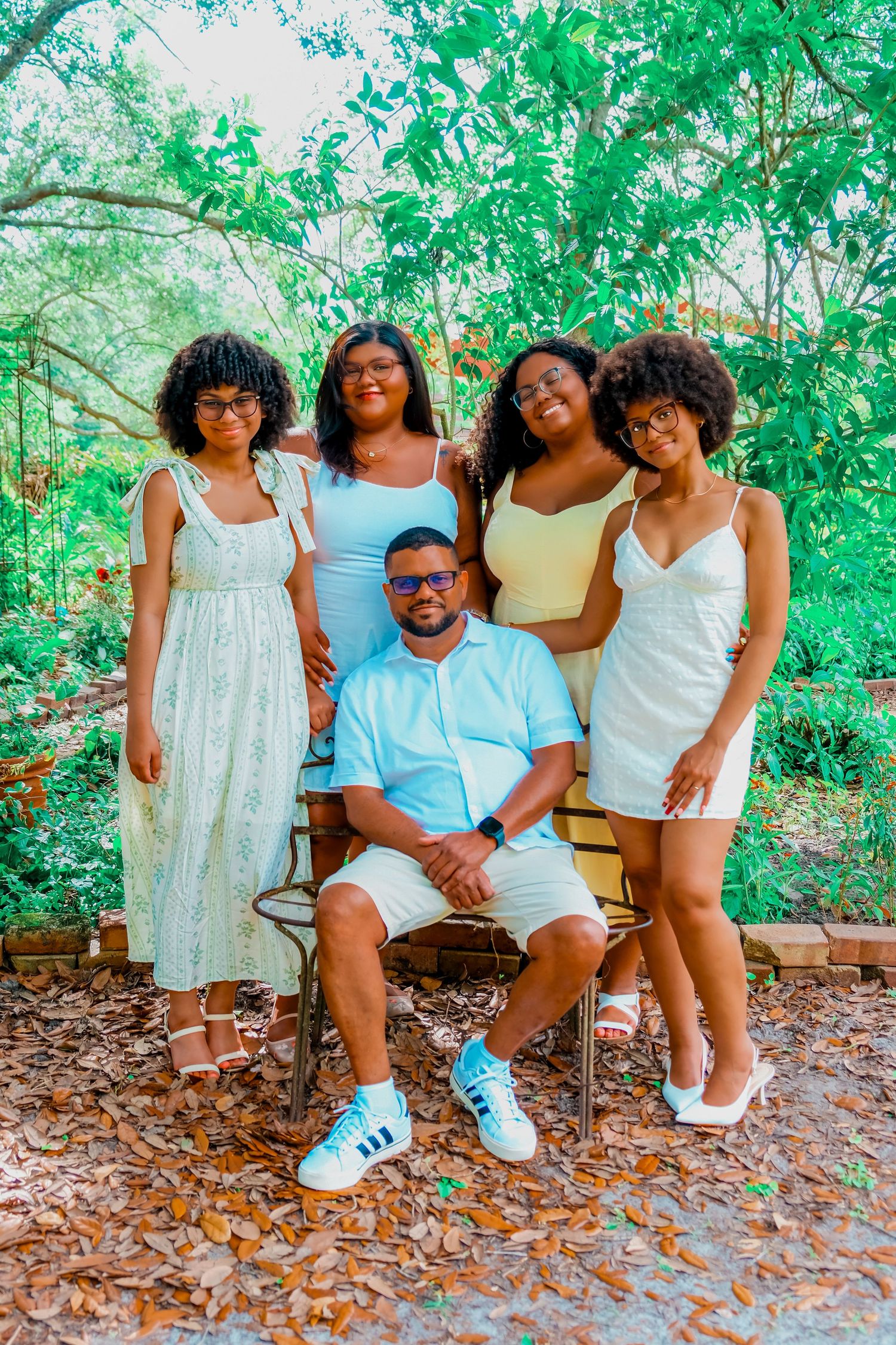 A family of five poses outdoors in a lush garden, with four women standing behind a seated man in casual summer attire.