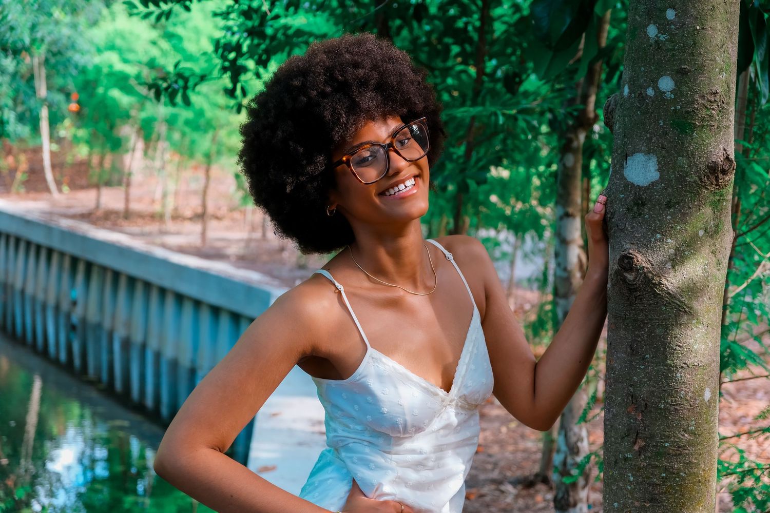 Smiling young Black woman with afro hair and glasses in a white dress leaning against a tree in a lush green park.