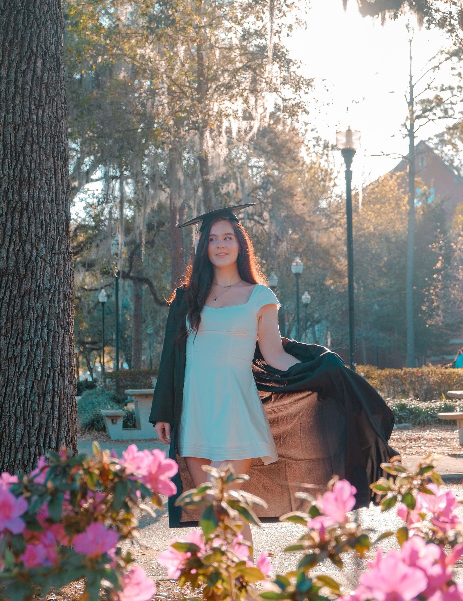 Young woman in graduation cap and gown poses among pink flowers and trees on a sunny college campus.