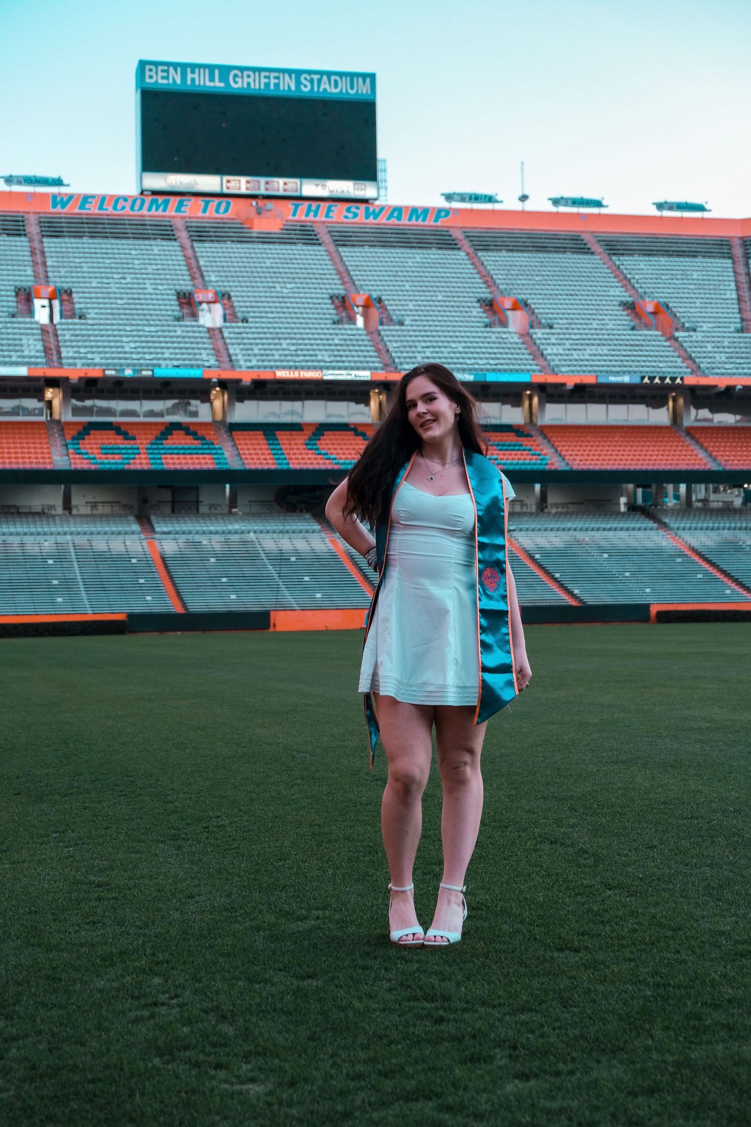 Young woman in white dress with teal graduation stole stands on field at Ben Hill Griffin Stadium, The Swamp.