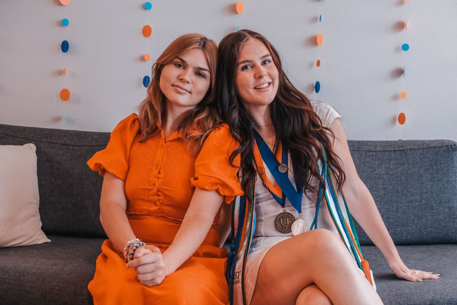 Two young women sitting together on a gray couch smiling, one wearing a bronze medal at a celebration.