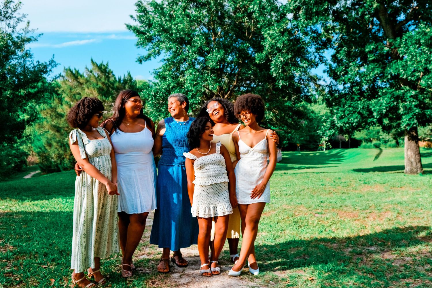 Five young women in summer dresses laugh together on a lush green park path surrounded by tall trees.