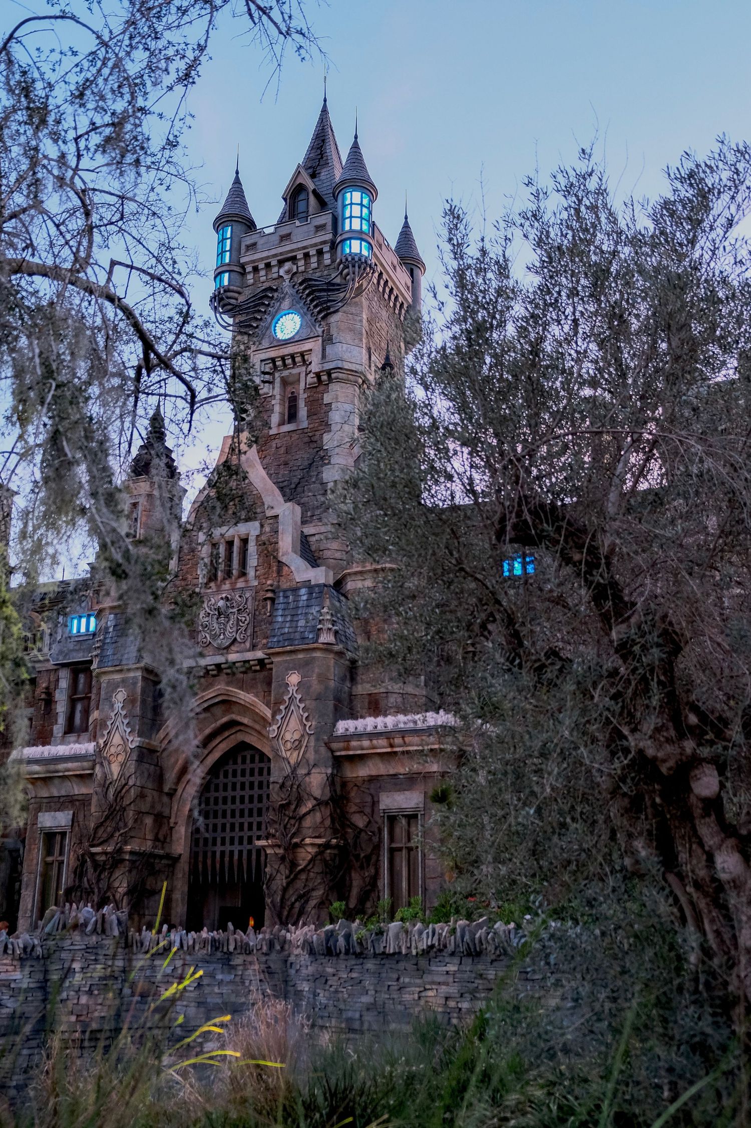 Gothic castle with clock tower and turrets surrounded by gnarled trees at dusk, glowing teal windows.