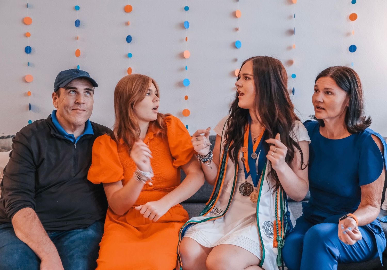 Four people sitting together laughing at a celebration, woman wearing medals and white dress, colorful garland backdrop.
