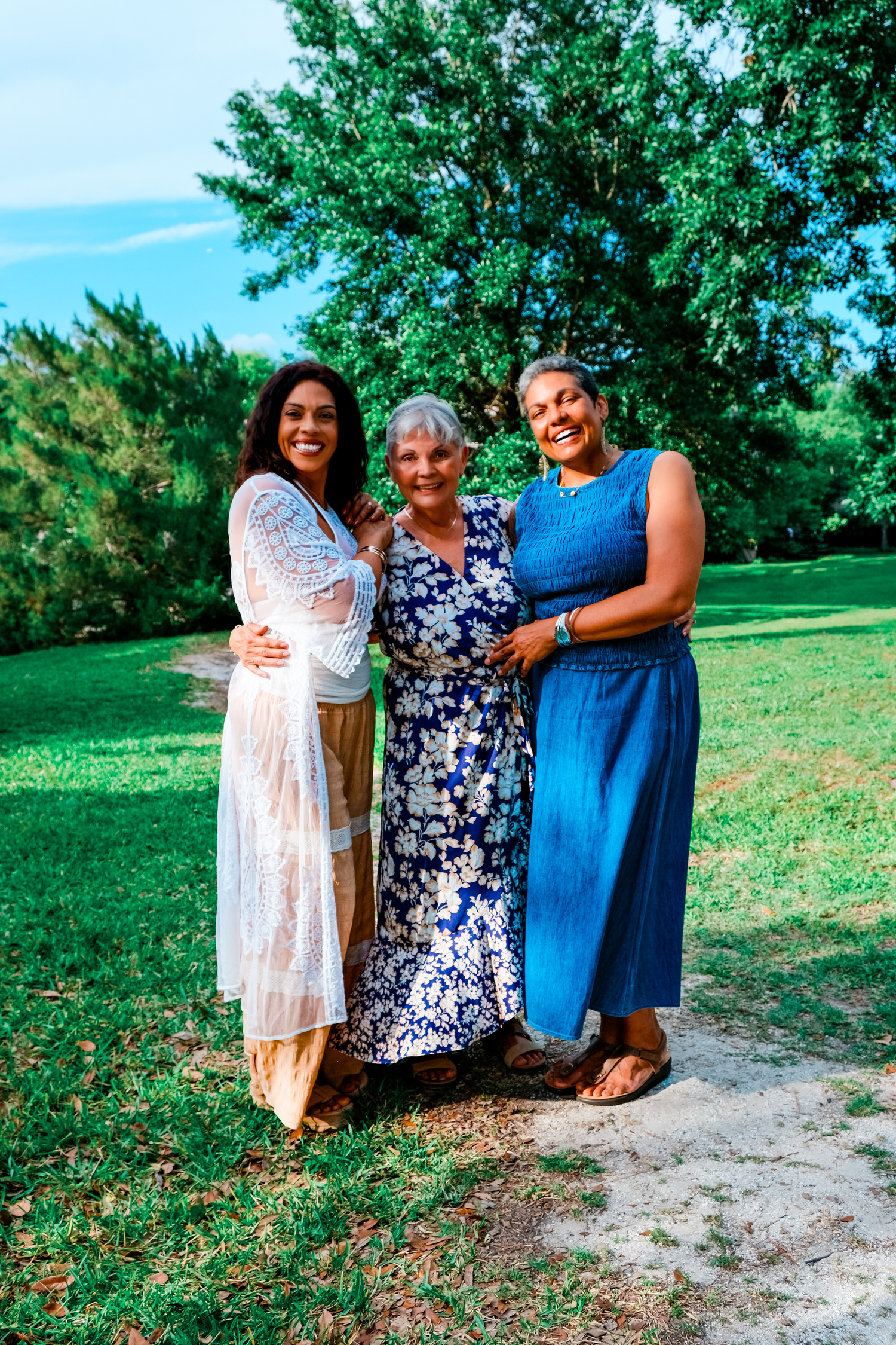 Three women smiling together outdoors in a lush green park, wearing floral and solid maxi dresses on a sunny day.