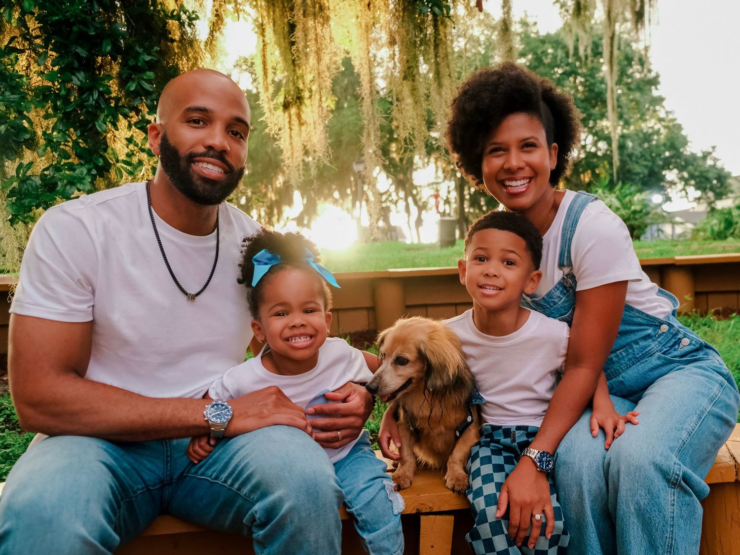 Happy Black family with two young children and a golden dog posing outdoors near lush green trees.