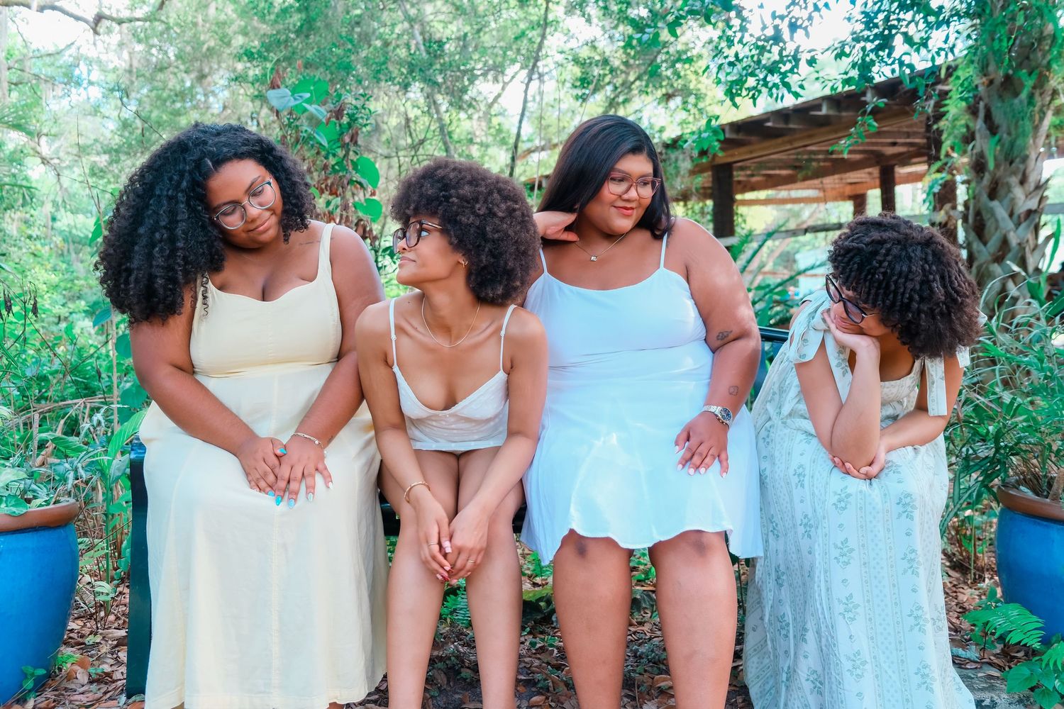 Four young Black women in pastel dresses sit together outdoors surrounded by lush green trees and plants.