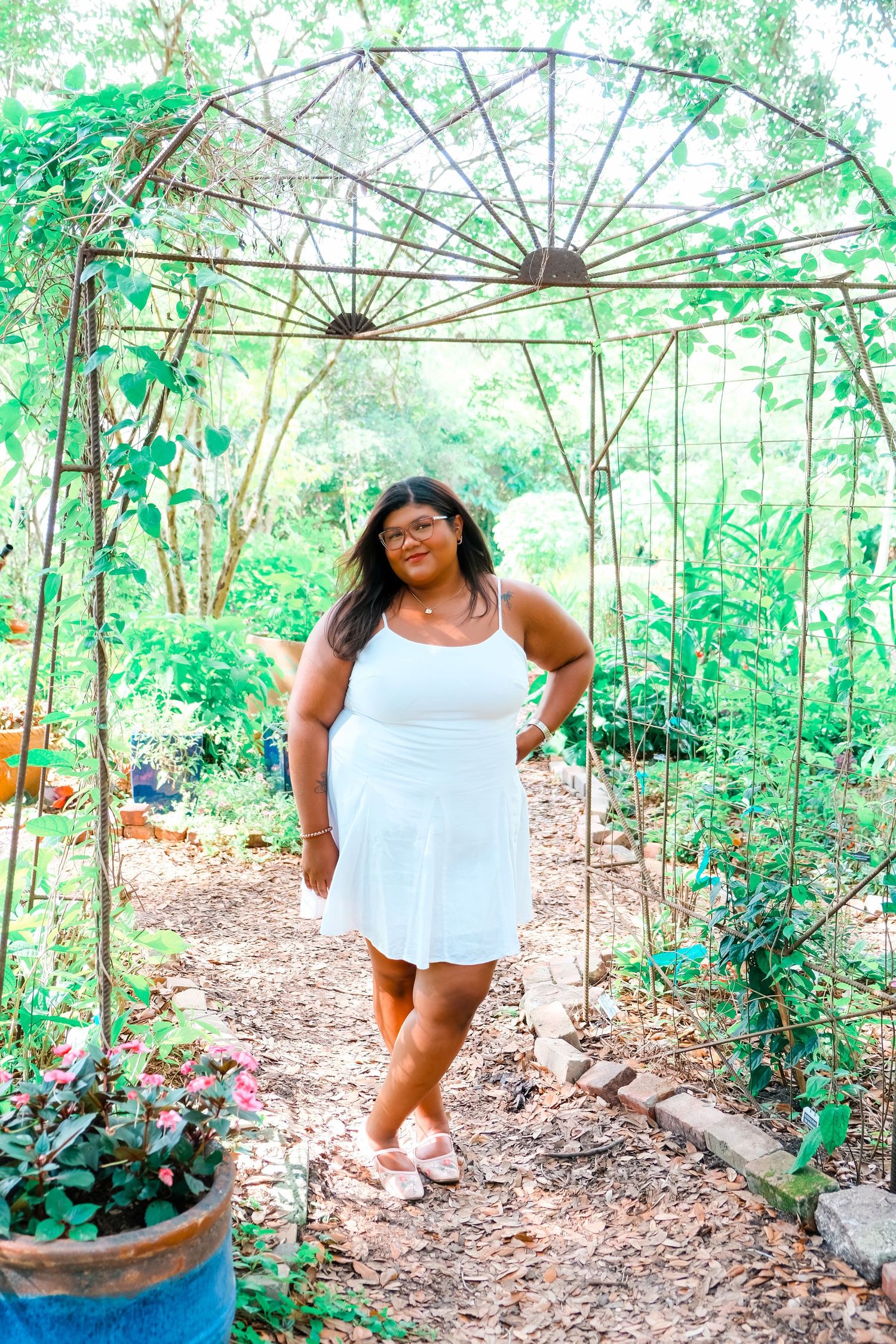 A young woman in a white dress poses under a rustic metal garden arbor surrounded by lush green plants.