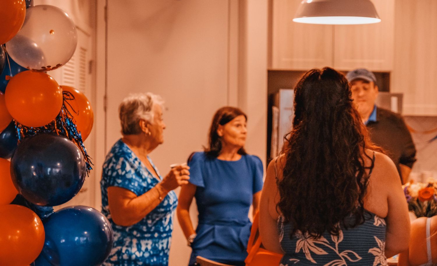 Group of people socializing at a party with blue and orange balloon decorations indoors.