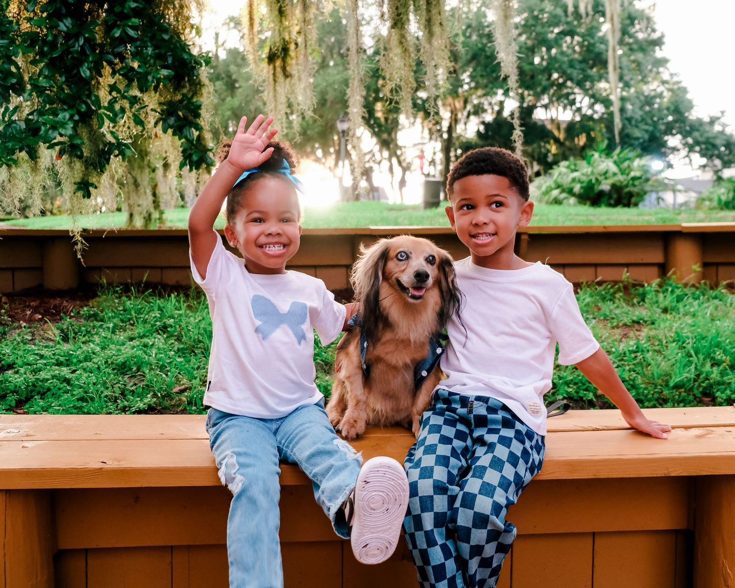 Two smiling children sit on a wooden bench with a dachshund dog between them in a lush green park setting.
