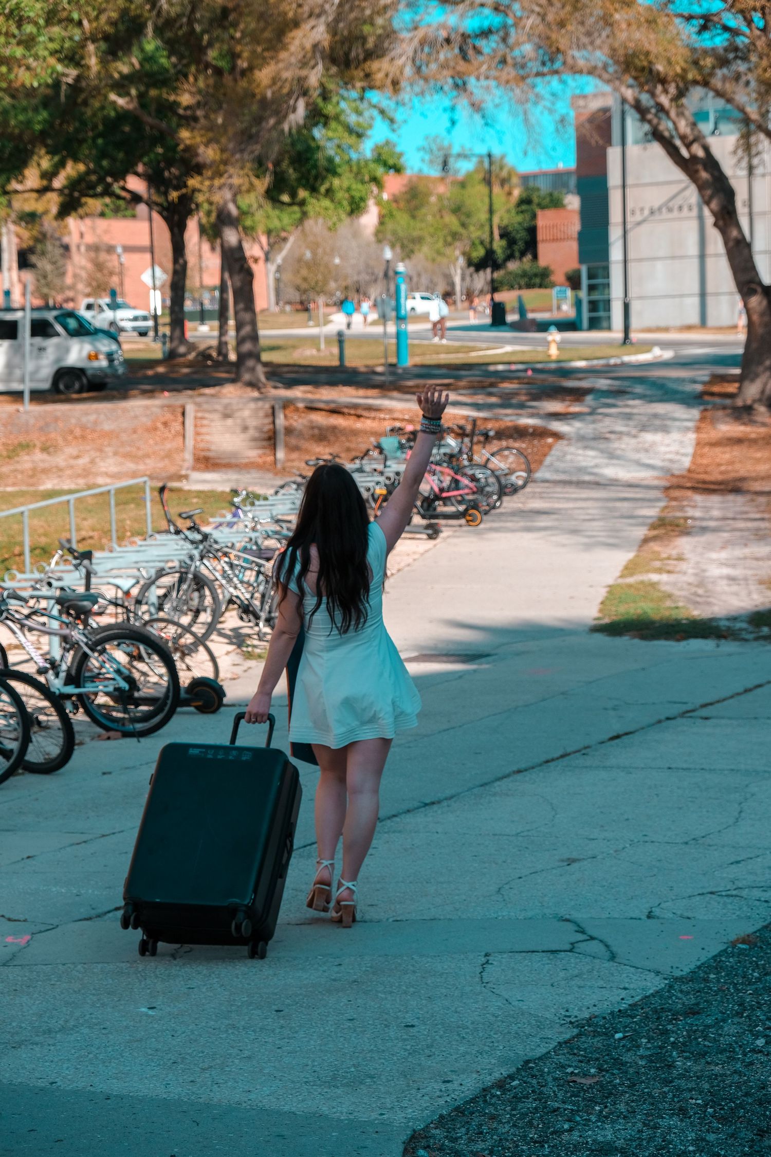 Female graduate in white dress walks away with suitcase, waving goodbye on a sunny college campus pathway.