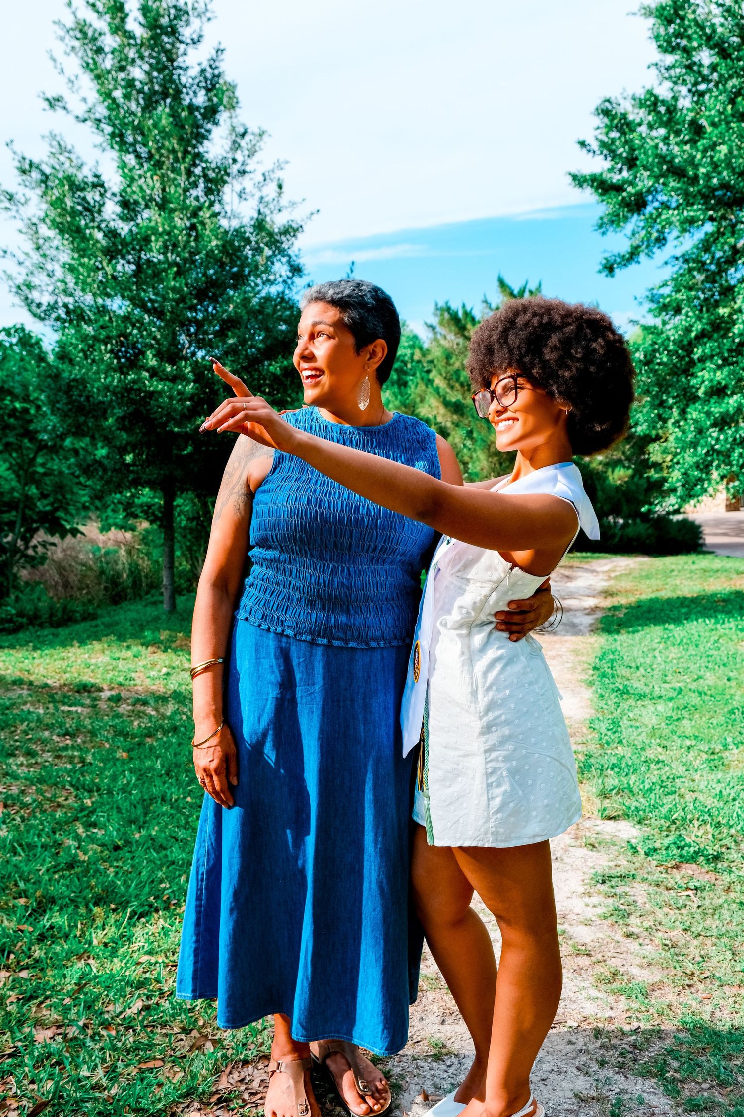 Two Black women smiling outdoors in a park, one in blue dress pointing while other in white dress embraces her.