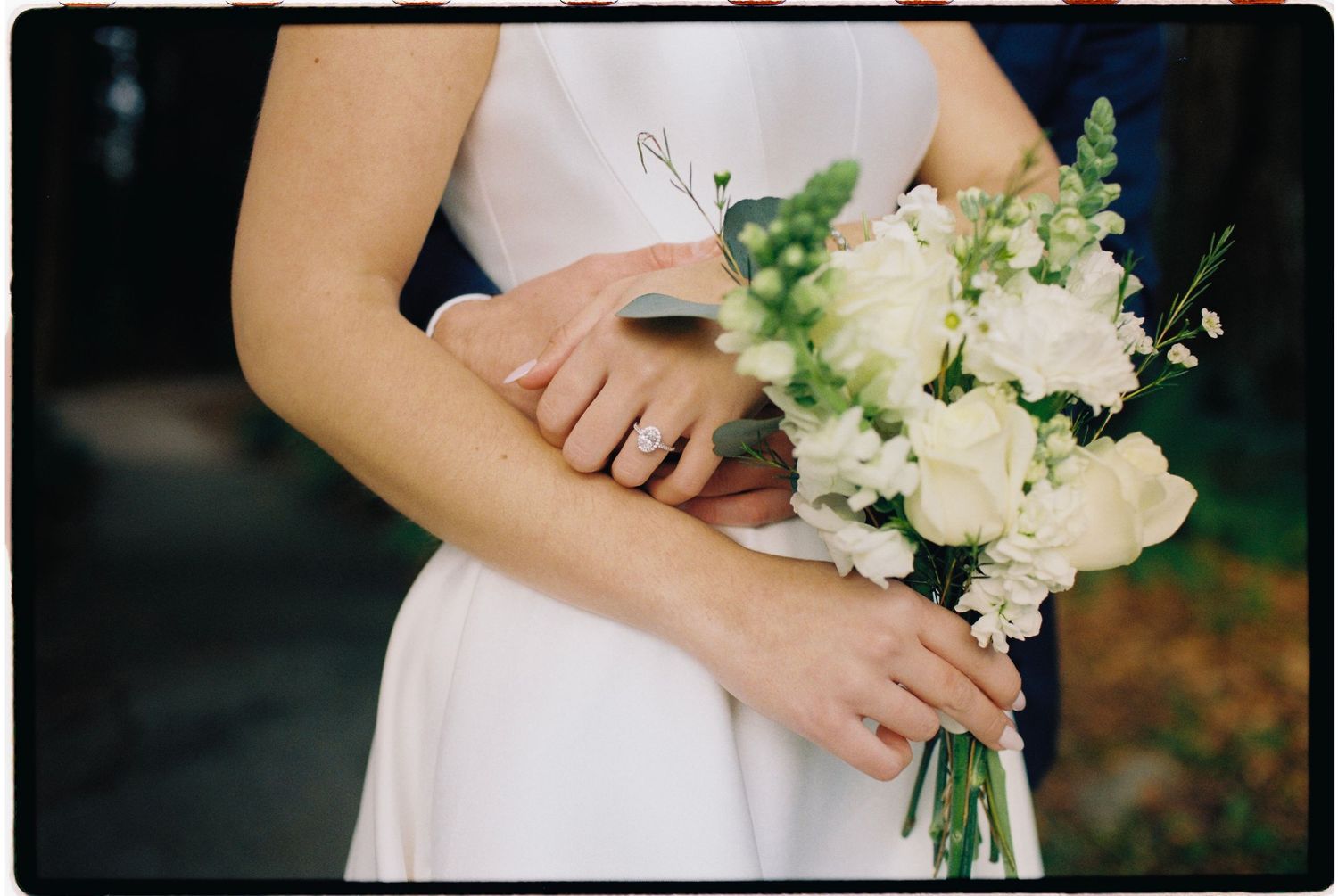 Close-up of bride holding white floral bouquet with groom embracing her from behind.