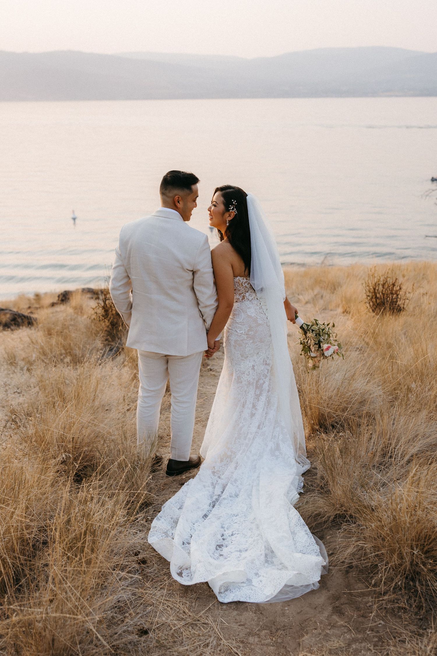 Bride and groom holding hands overlooking serene lake at golden hour during outdoor wedding.