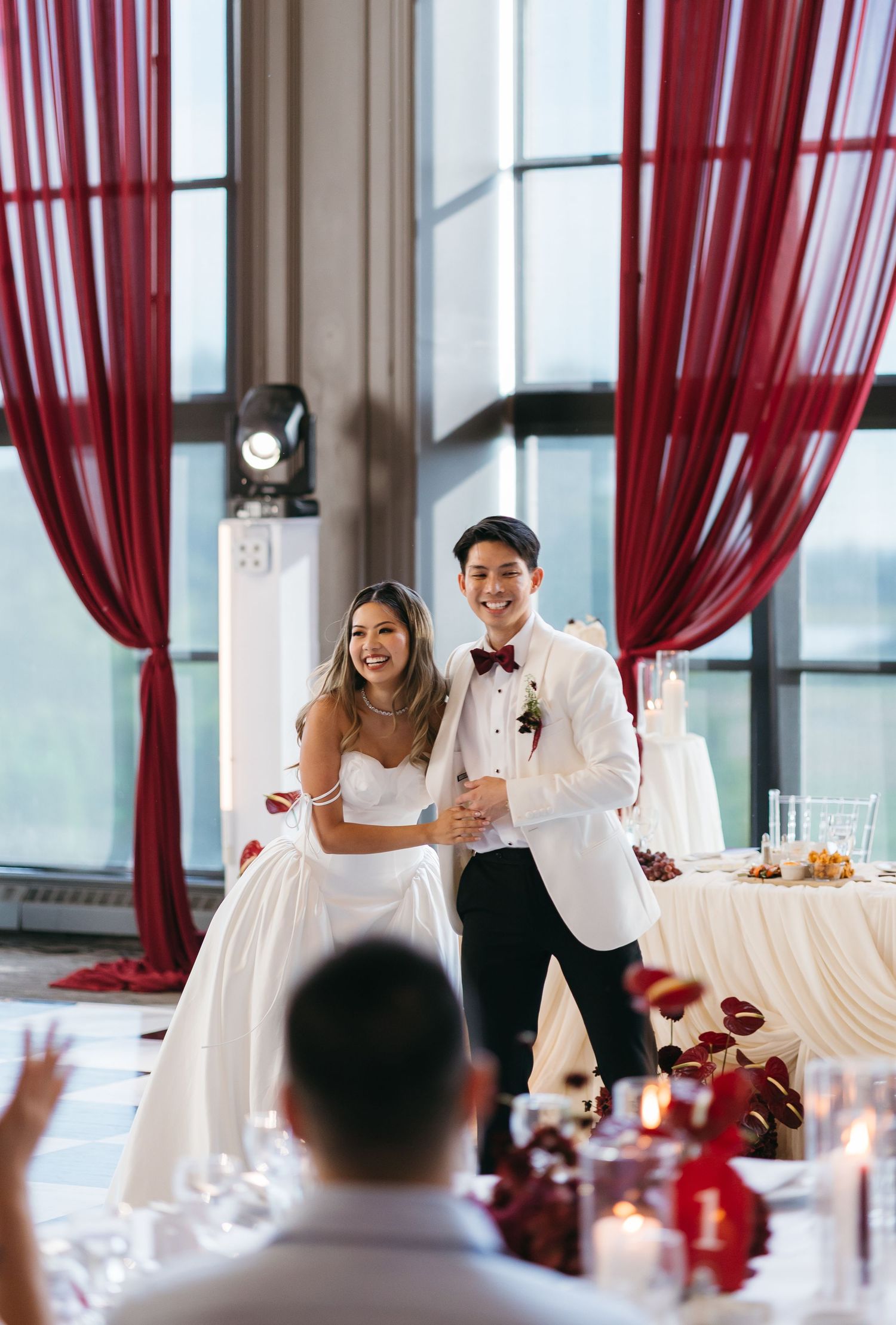 Bride in white gown and groom in white tuxedo smiling at wedding reception with red drape decor.