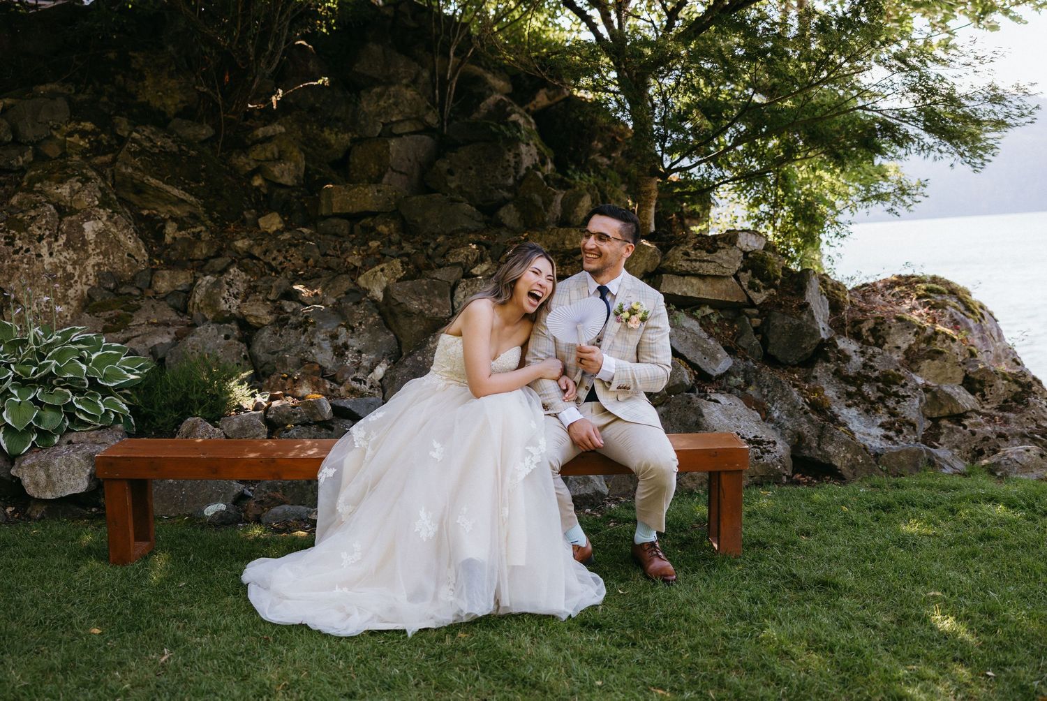 Laughing bride and groom sit on wooden bench beside stone wall and waterfront garden setting.